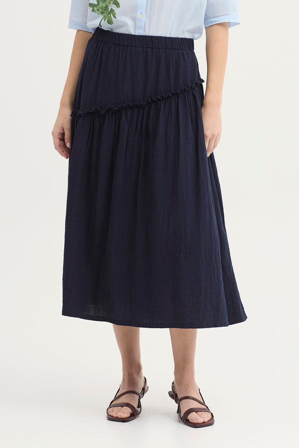 Close up of women wearing a light blue short-sleeve blouse and a dark navy tiered midi skirt against a plain light background.
