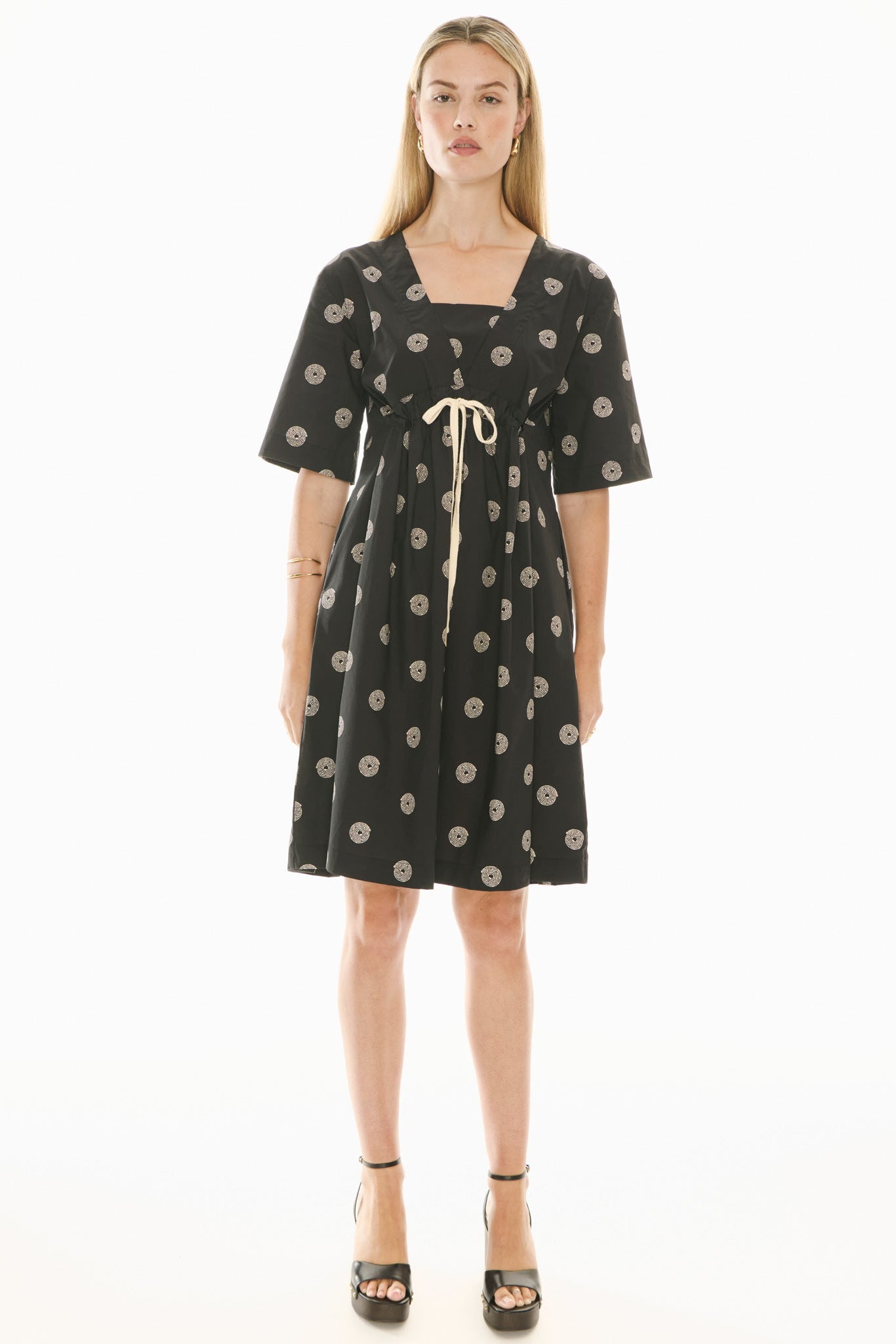 Woman wearing a short sleeve black dress with beige pattern and beige waist tie on a white background