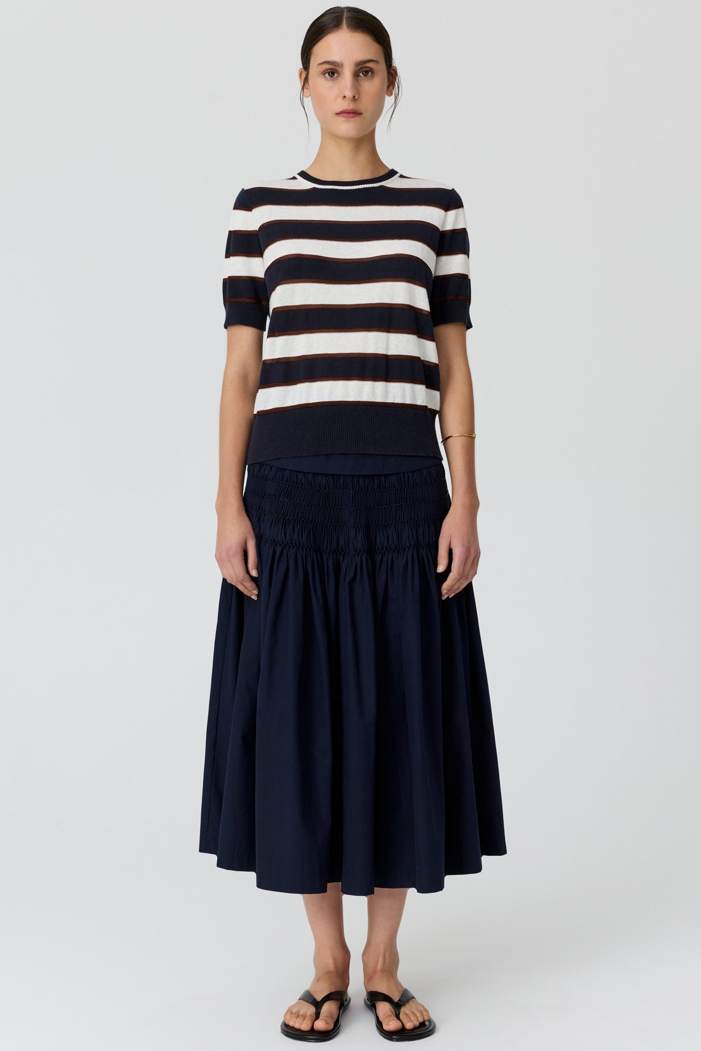 A woman wearing a navy and cream horizontally striped short-sleeve knit top and a navy tiered midi skirt, paired with black flat sandals, against a light grey studio background.