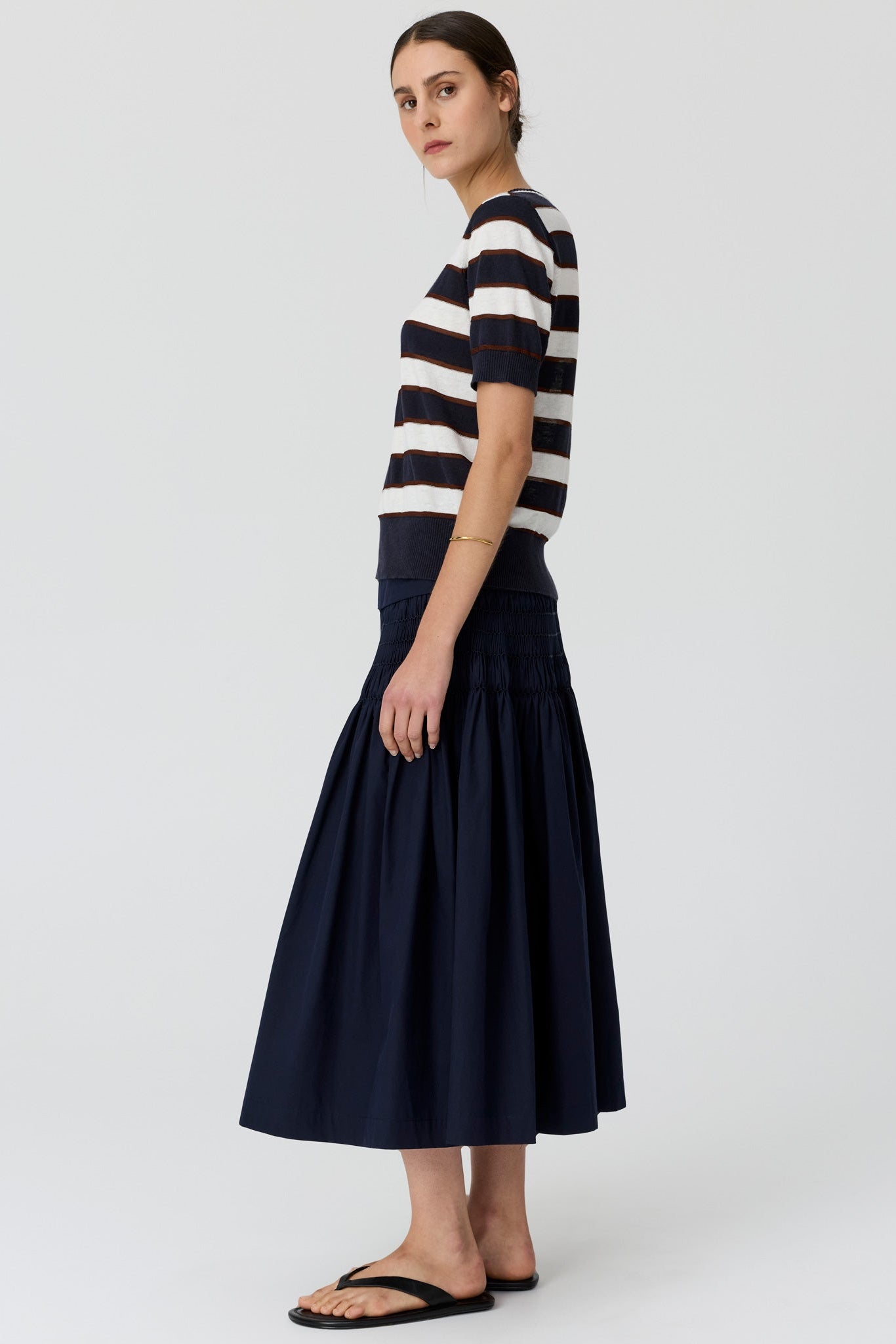 A woman viewed from the side wearing a navy and cream horizontally striped short-sleeve knit top and a navy tiered midi skirt, paired with black flat sandals, against a light grey studio background.