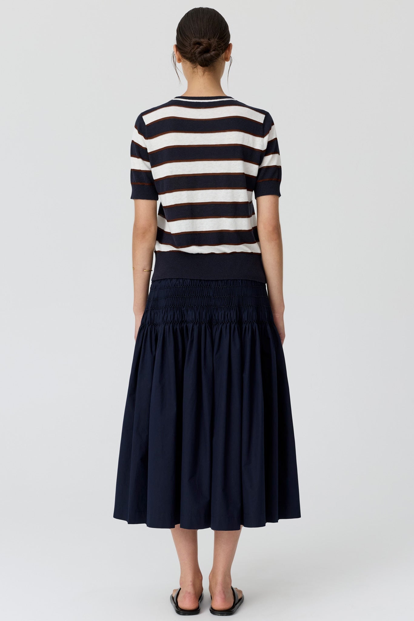 A woman viewed from the back wearing a navy and cream horizontally striped short-sleeve knit top and a navy tiered midi skirt, paired with black flat sandals, against a light grey studio background.