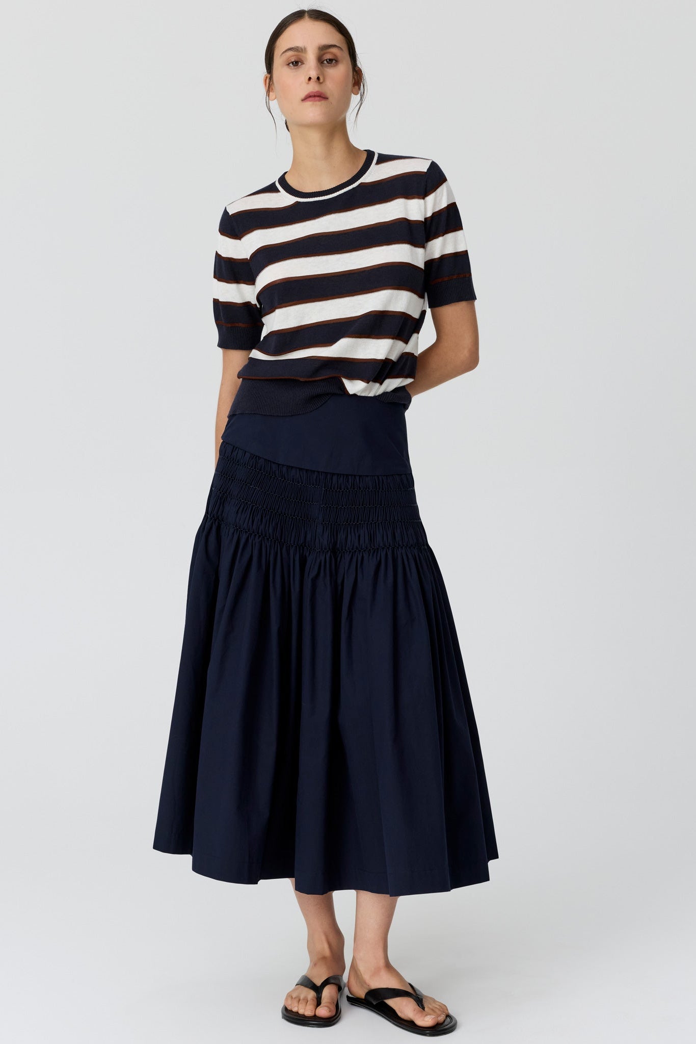 A woman wearing a navy and cream horizontally striped short-sleeve knit top and a navy tiered midi skirt, paired with black flat sandals, against a light grey studio background.