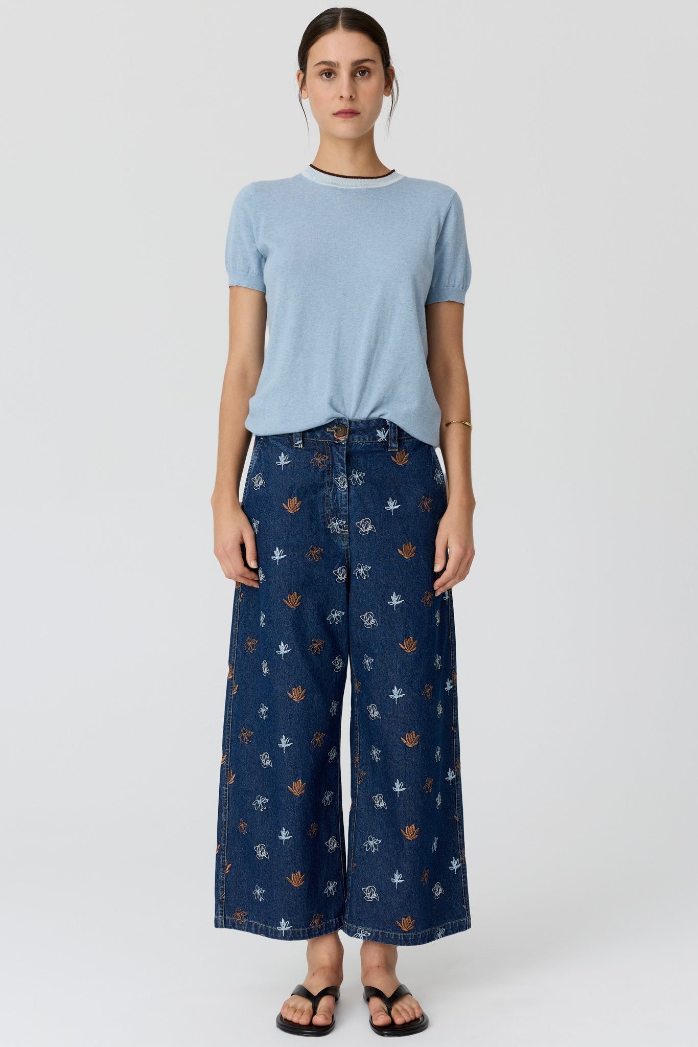 A model stands against a light gray studio background wearing a light blue short-sleeve t-shirt and dark blue wide-leg pants with a small floral pattern.