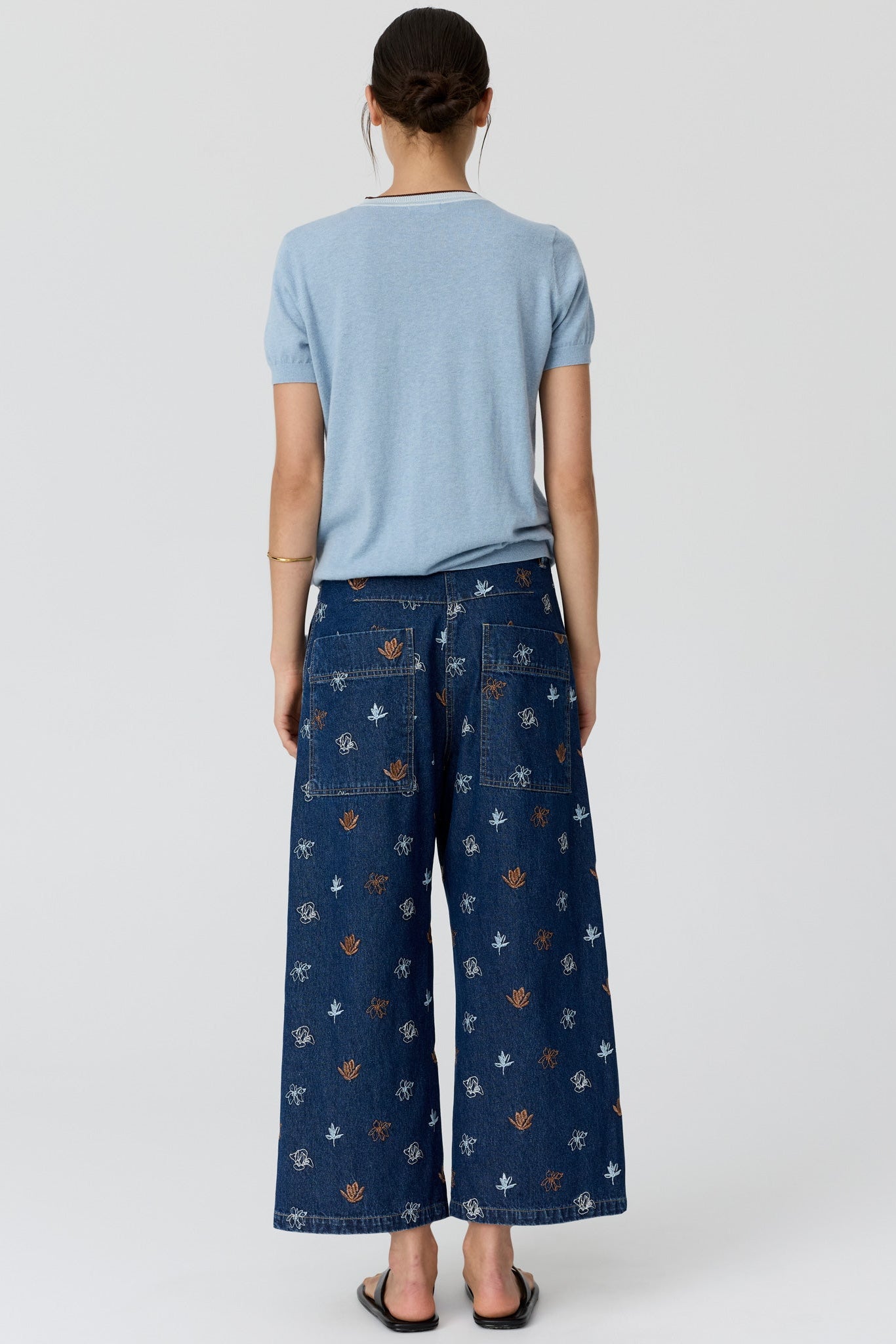 A model shown from the back wearing a light blue short-sleeve t-shirt and dark blue wide-leg denim pants with a small orange and white pattern, set against a plain light gray background.
