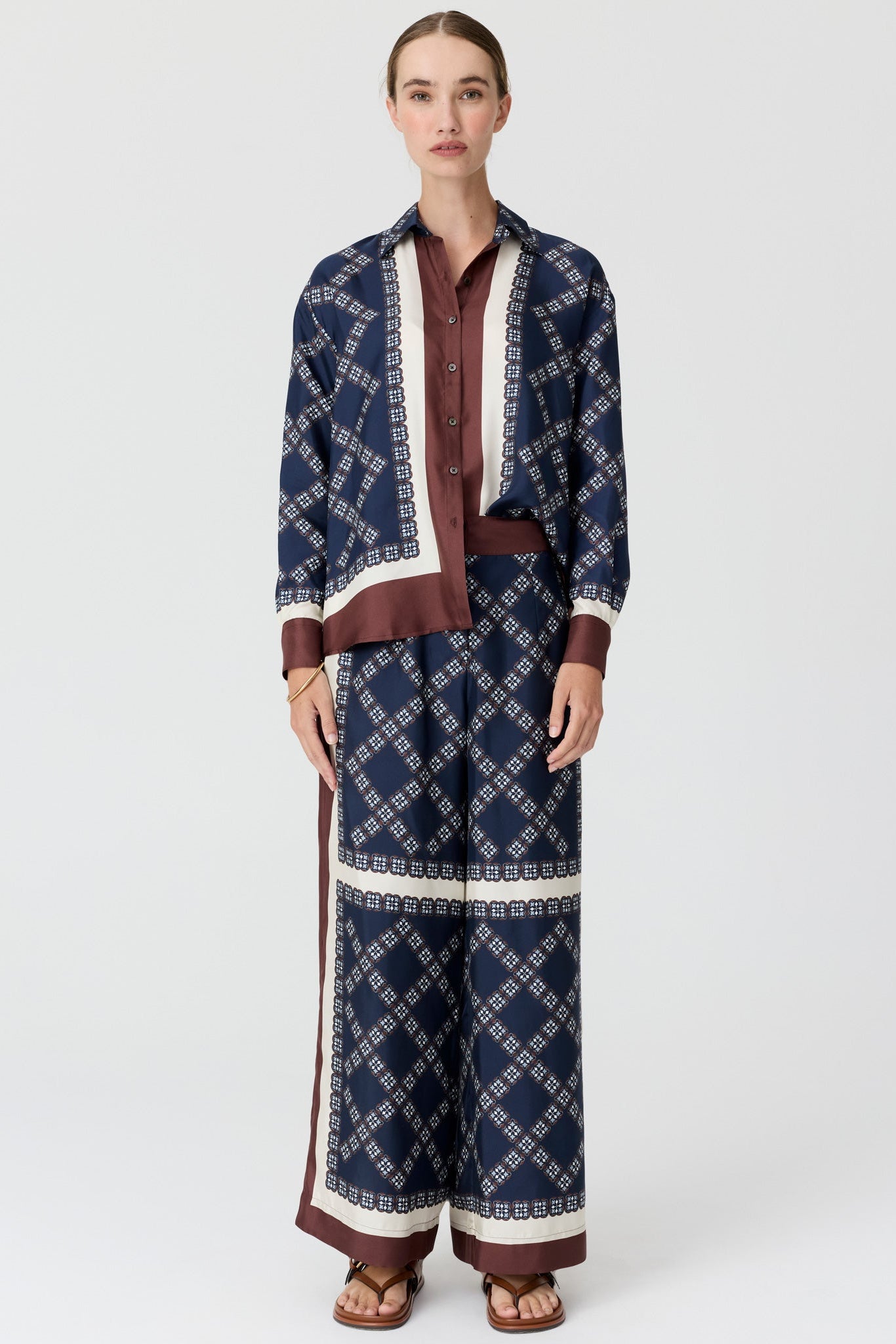 A model wearing a matching navy blue patterned long-sleeve shirt and wide-leg trousers set with brown and cream details, on a light grey studio background.