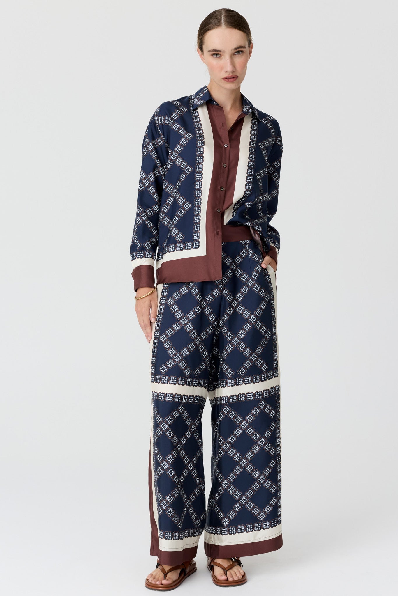 A model wearing a matching navy blue patterned long-sleeve shirt and wide-leg trousers set with brown and cream details, on a light grey studio background.