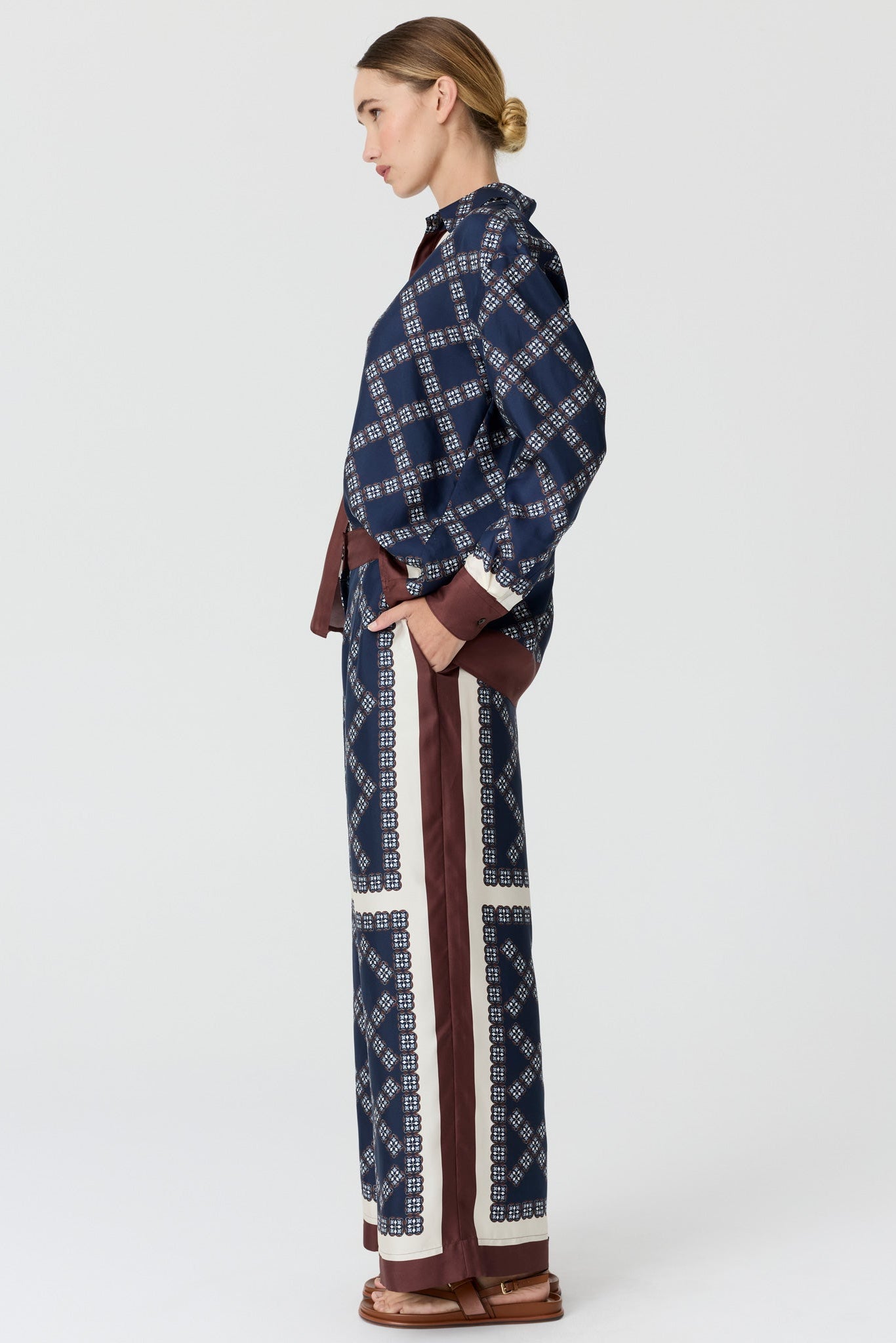 A model side view wearing a matching navy blue patterned long-sleeve shirt and wide-leg trousers set with brown and cream details, on a light grey studio background.