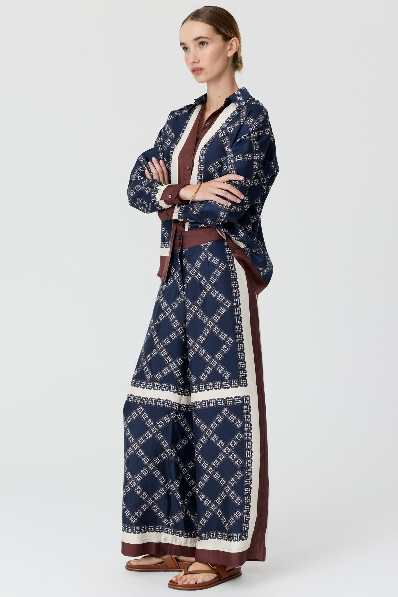 A model side on wearing a matching navy blue patterned long-sleeve shirt and wide-leg trousers set with brown and cream details, on a light grey studio background.