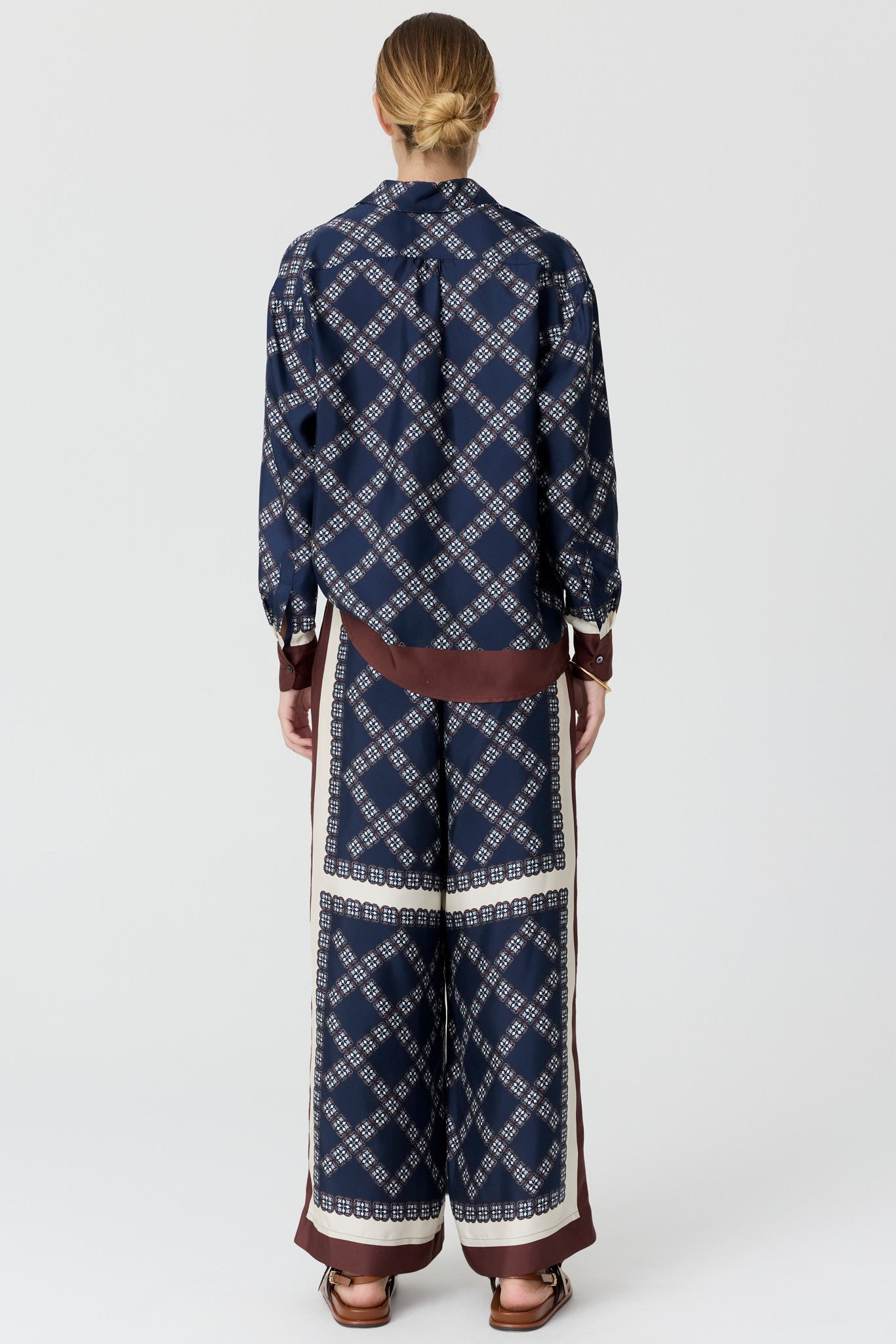 A model viewed from the back wearing a matching navy blue patterned long-sleeve shirt and wide-leg trousers set with brown and cream details, on a light grey studio background.