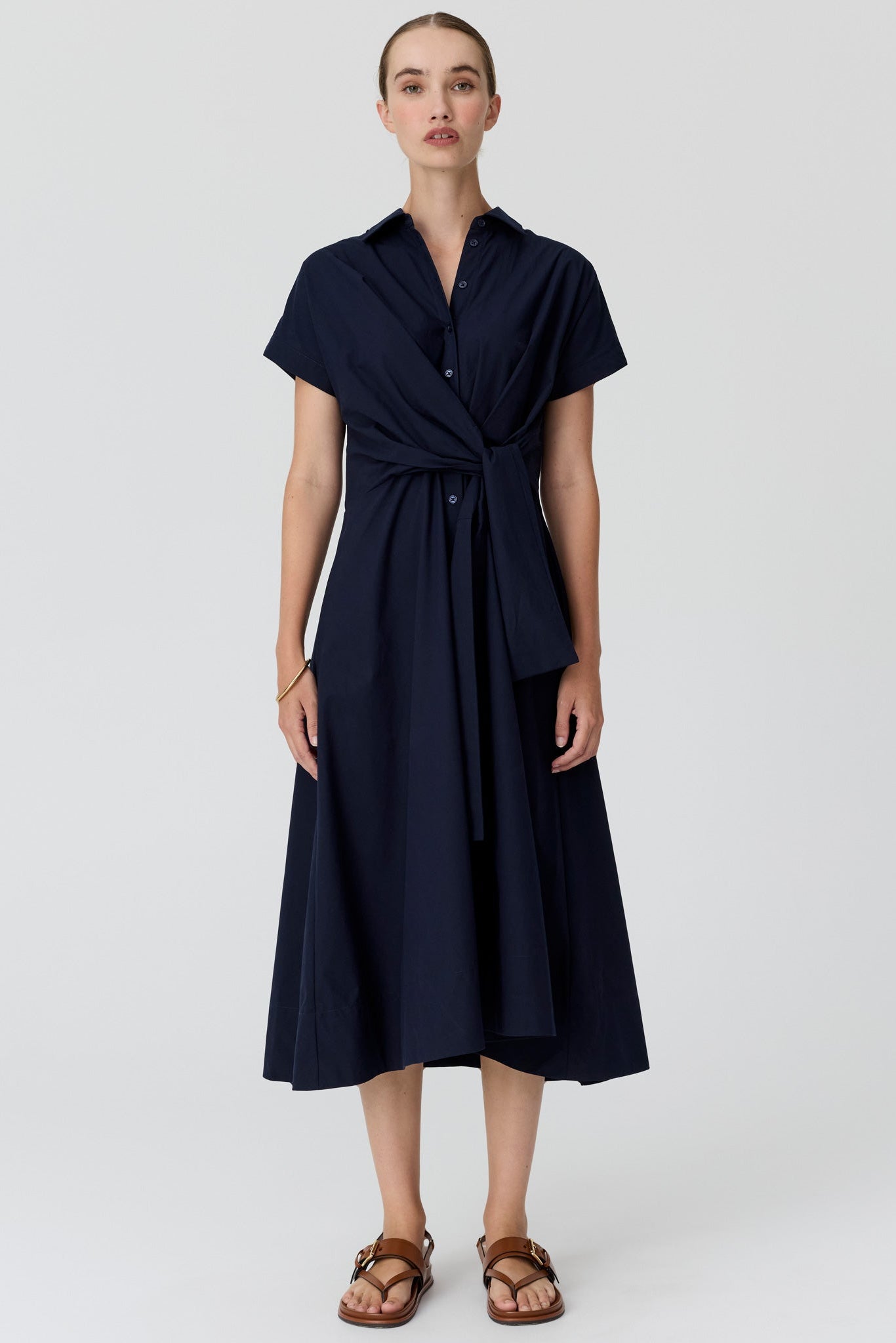 A woman wearing a dark navy midi dress with short sleeves and a collared neckline, paired with brown sandals, against a light grey studio background.