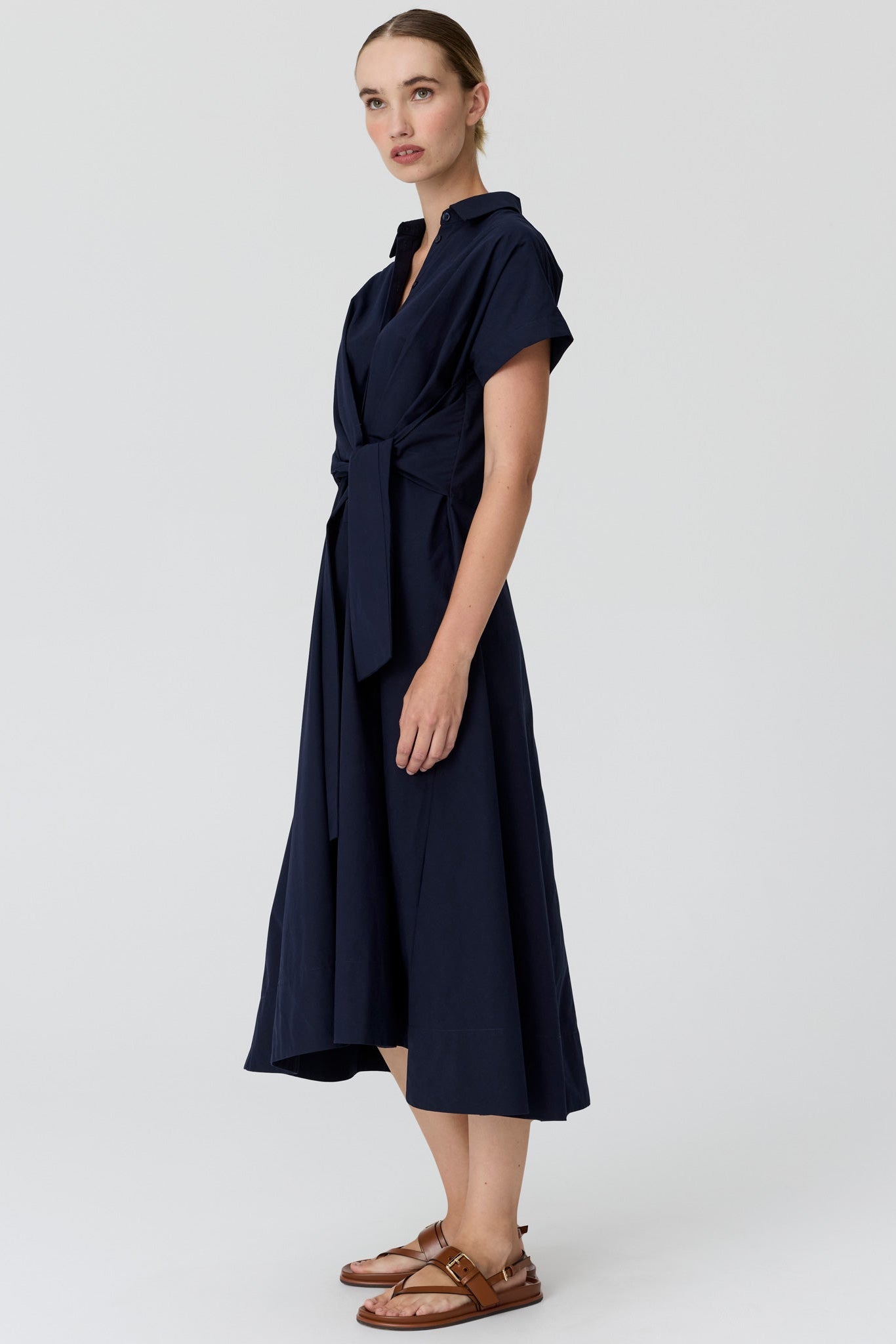 A woman viewed from the side wearing a dark navy midi dress with short sleeves and a collared neckline, paired with brown sandals, against a light grey studio background.