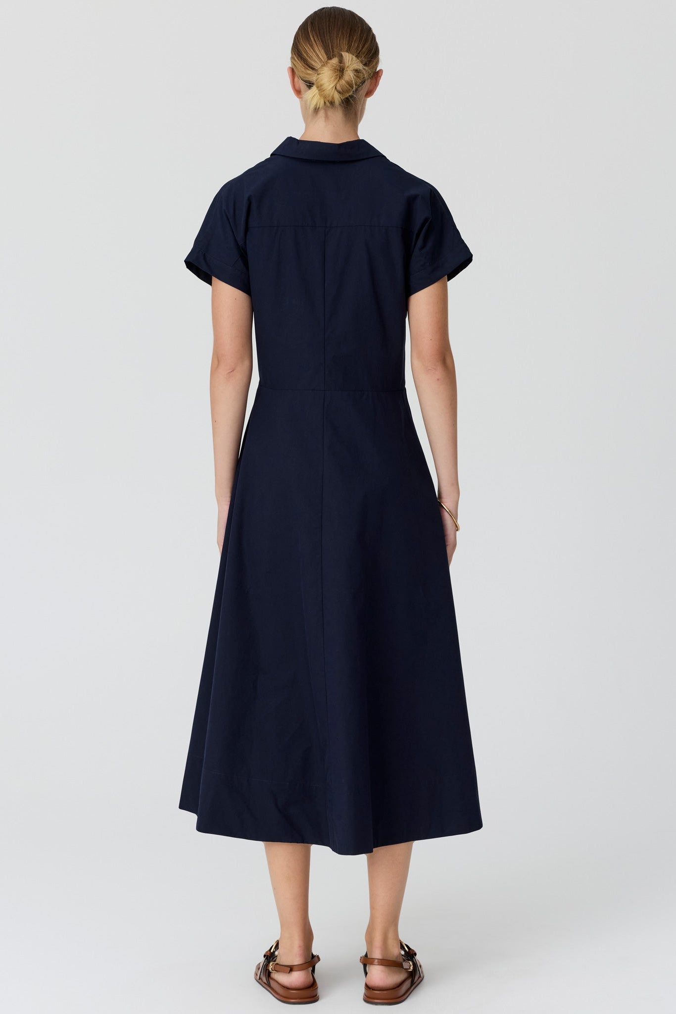 A woman viewed from the back wearing a dark navy midi dress with short sleeves and a collared neckline, paired with brown sandals, against a light grey studio background.