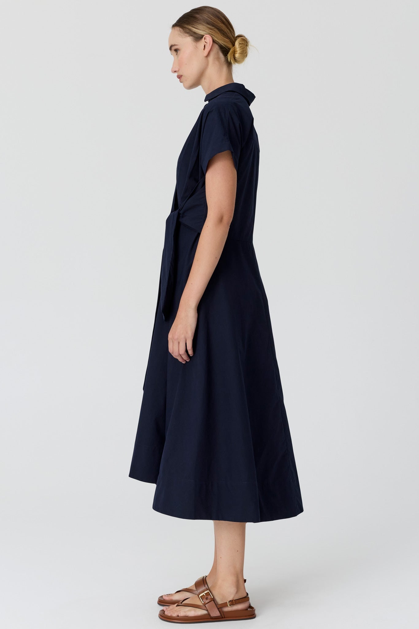 A woman viewed from the side wearing a dark navy midi dress with short sleeves and a collared neckline, paired with brown sandals, against a light grey studio background.