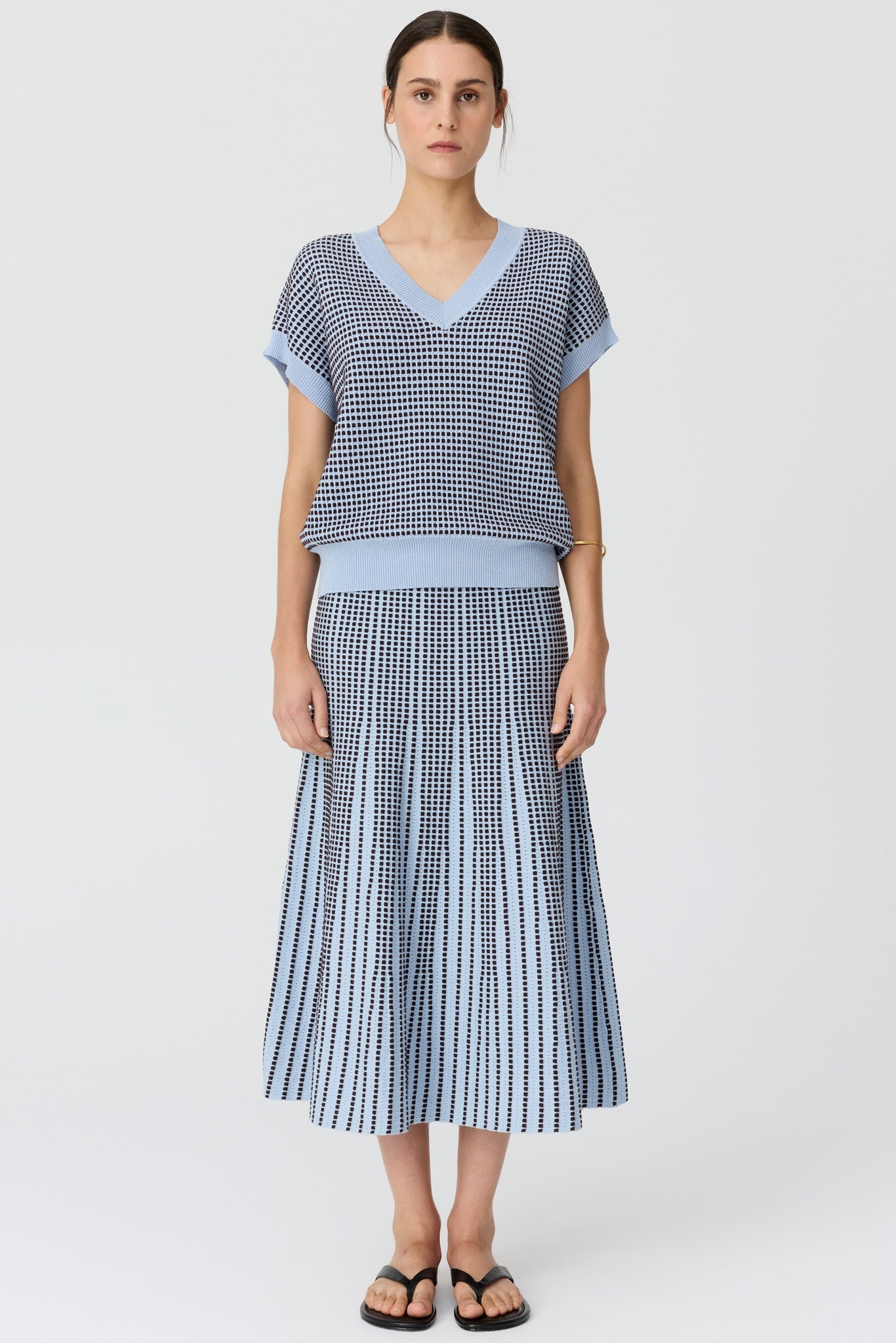 Women wearing a v neck short sleeve knit top in blue and chocolate pattern with a matching skirt and black sandals on a grey studio background. 