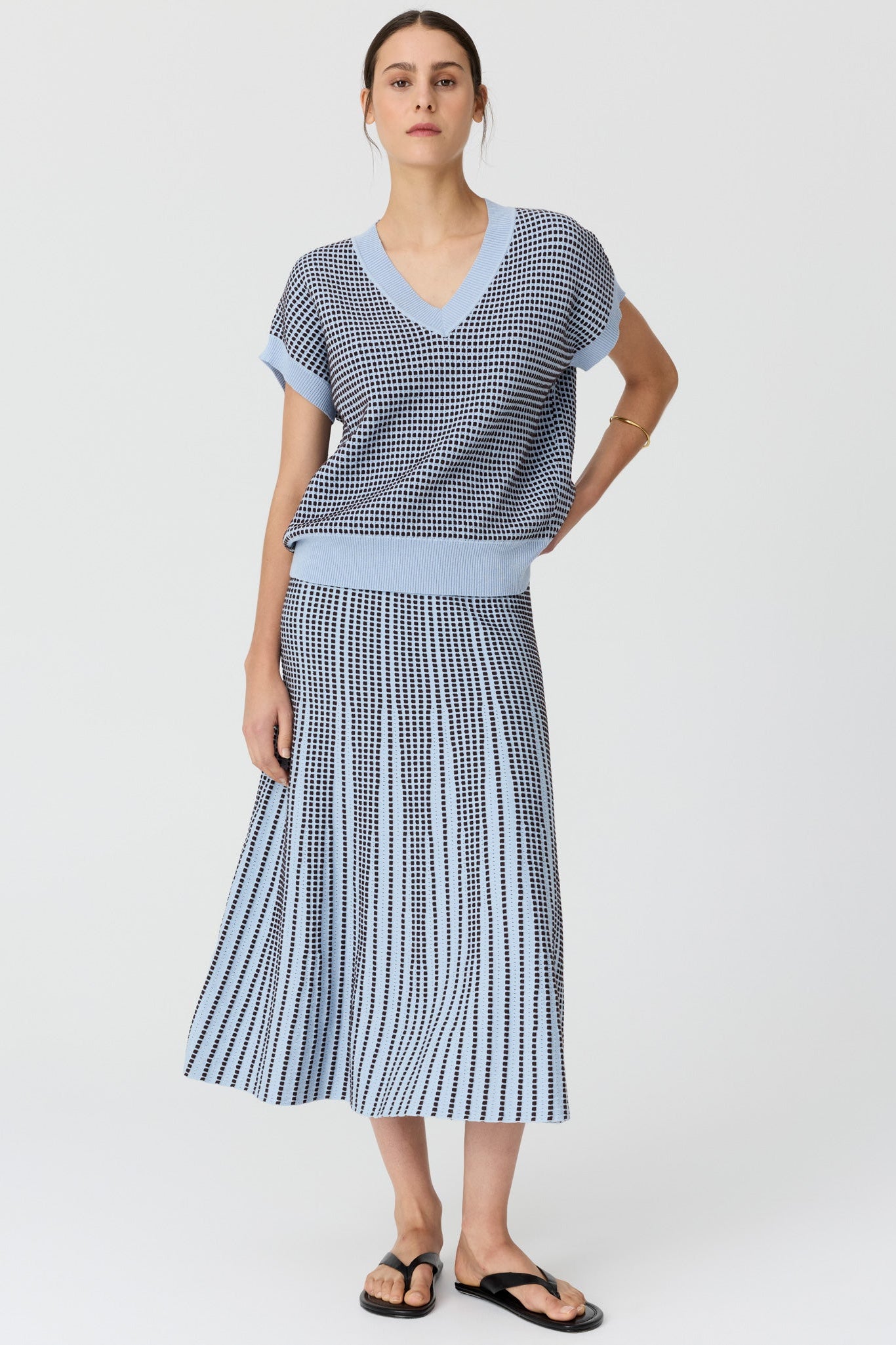 Women posing wearing a v neck short sleeve knit top in blue and chocolate pattern with a matching skirt and black sandals on a grey studio background. 
