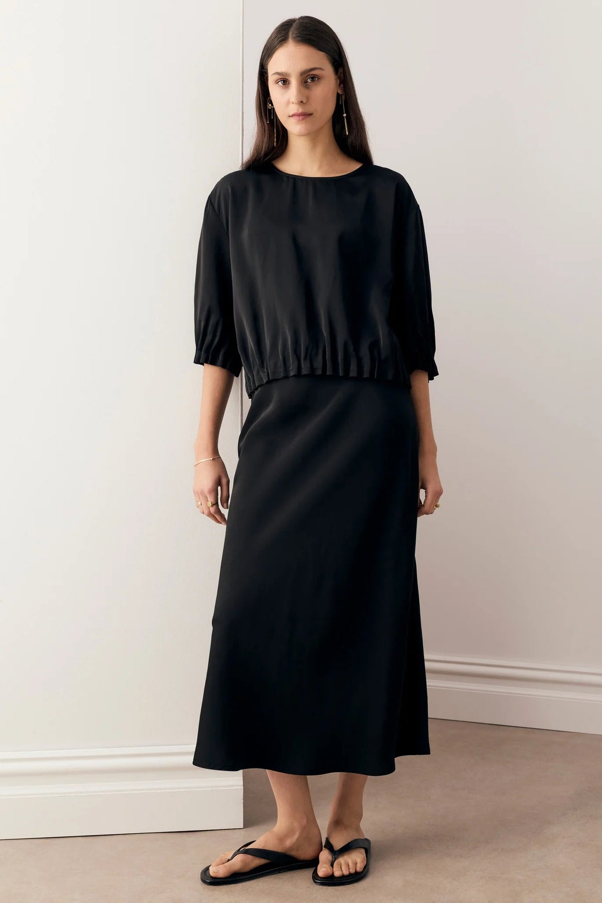 A woman wearing a black top with voluminous elbow-length puff sleeves and matching skirt and black flat sandals, photographed against a light studio background.
