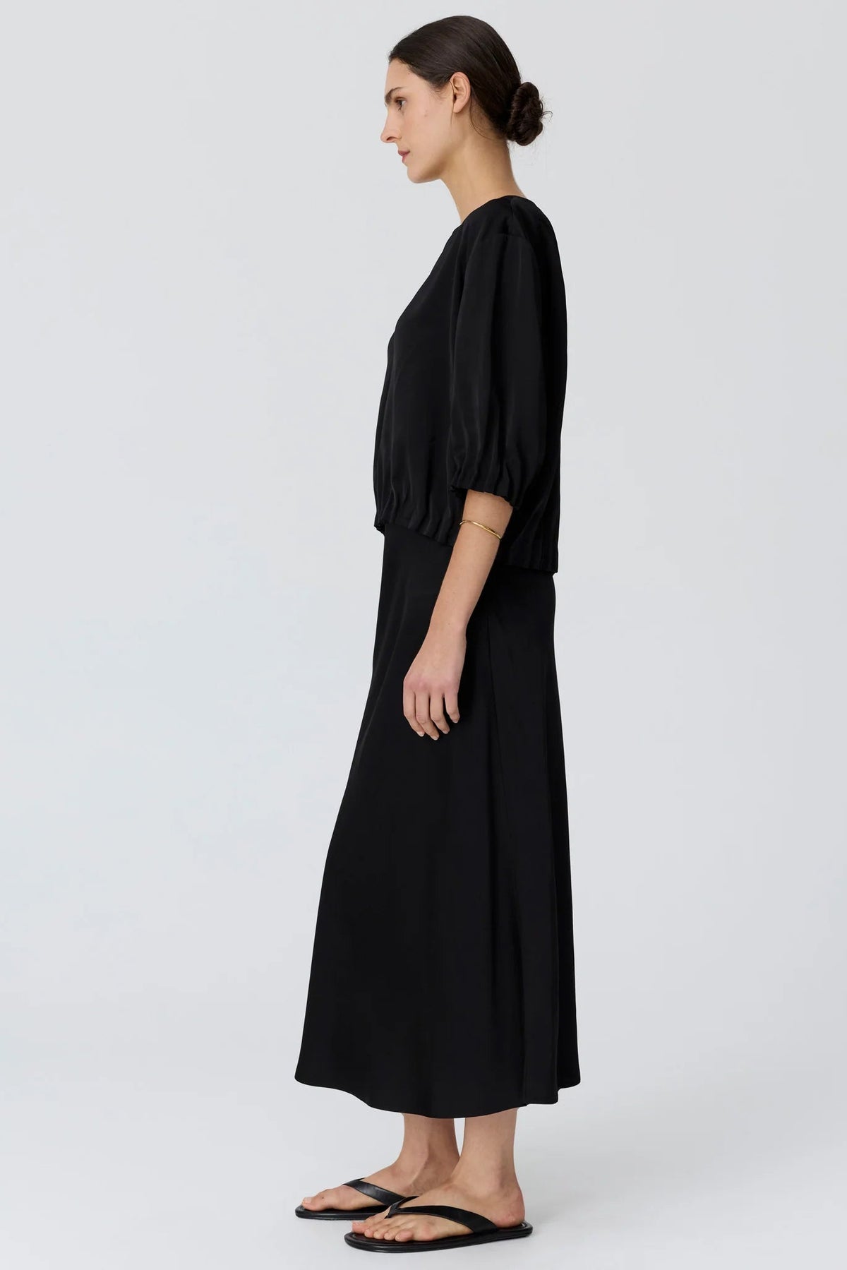 A woman in profile wearing a black top with voluminous elbow-length puff sleeves and matching skirt and black flat sandals, photographed against a light studio background.