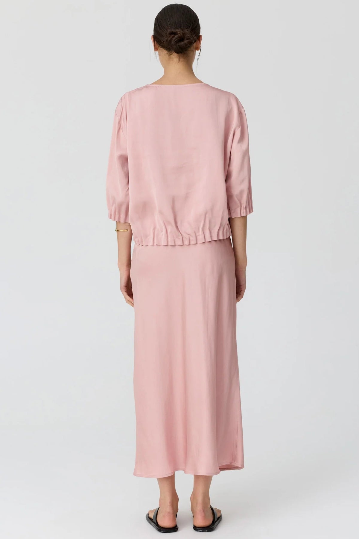 A woman shown from the back modeling a dusty pink two-piece set consisting of a short puff-sleeve top and a matching midi skirt, paired with black flip-flop sandals against a light gray studio background.  