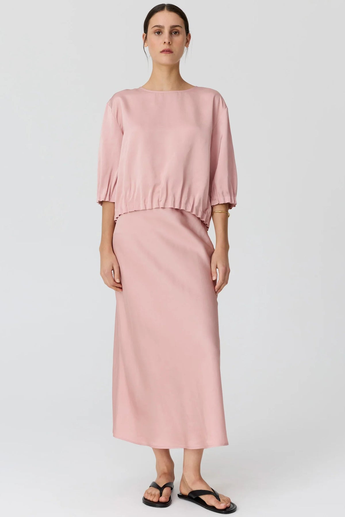 A woman modeling a dusty pink two-piece set consisting of a short puff-sleeve top and a matching midi skirt, paired with black flip-flop sandals against a light gray studio background.