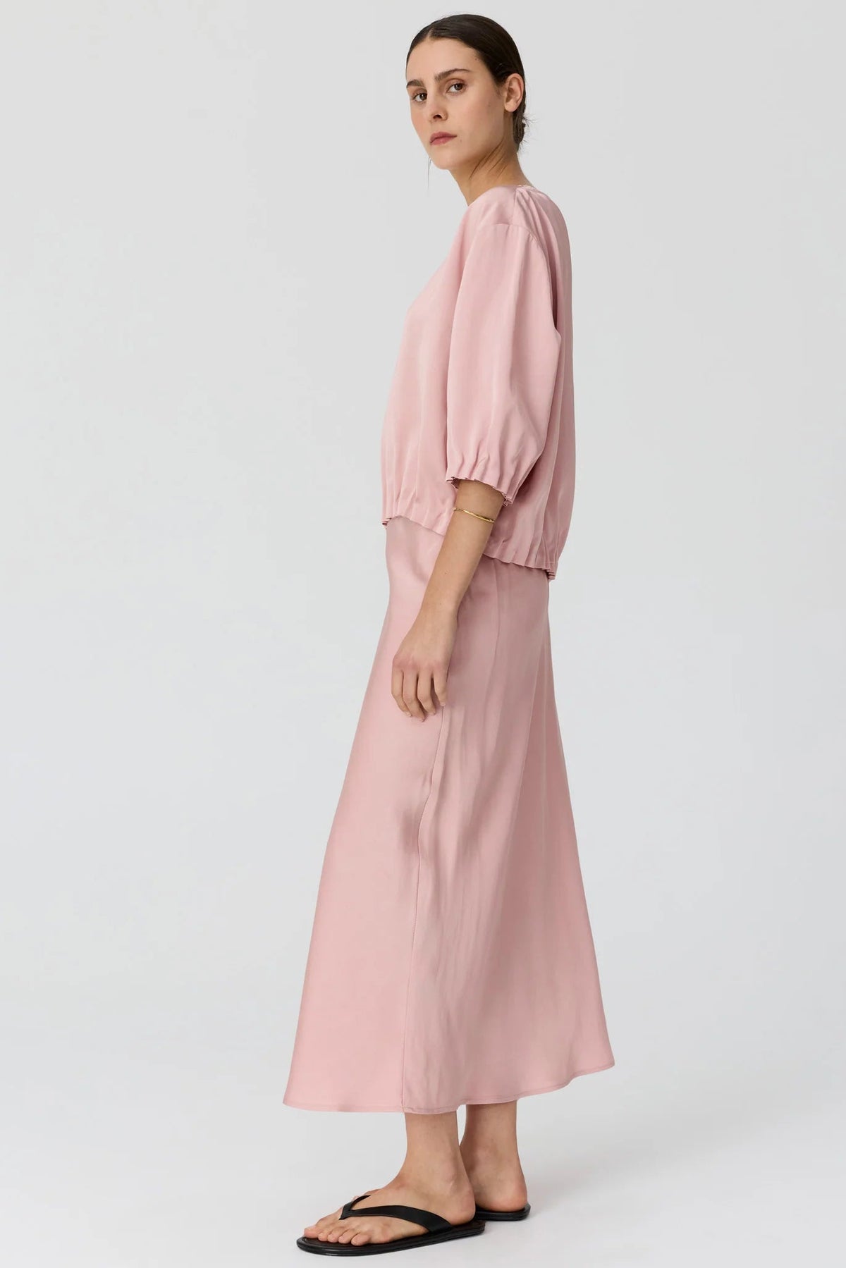 A woman modeling a dusty pink two-piece set consisting of a short puff-sleeve top and a matching midi skirt, paired with black flip-flop sandals against a light gray studio background.