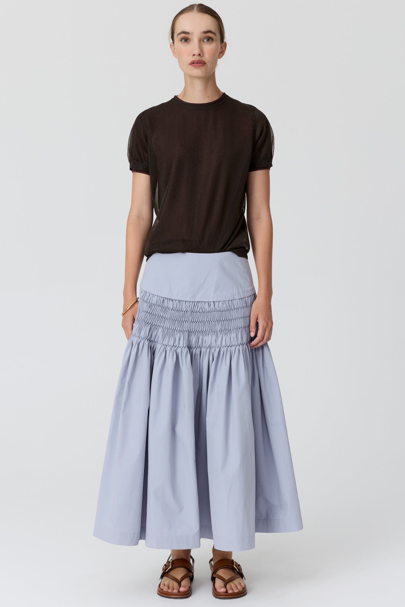 A model stands against a light gray studio background wearing a dark brown short-sleeve crewneck t-shirt, a light blue-gray smocked maxi skirt, and brown leather sandals.