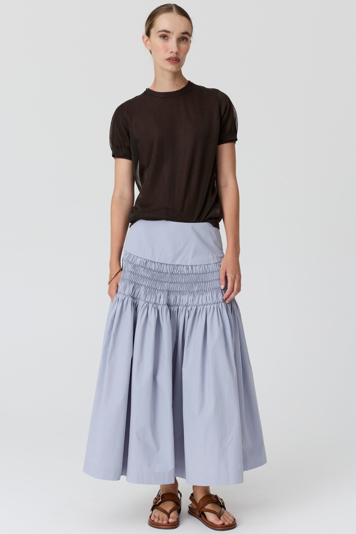 A model stands against a light gray studio background wearing a dark brown short-sleeve t-shirt, a light blue-gray smocked maxi skirt, and brown leather sandals.