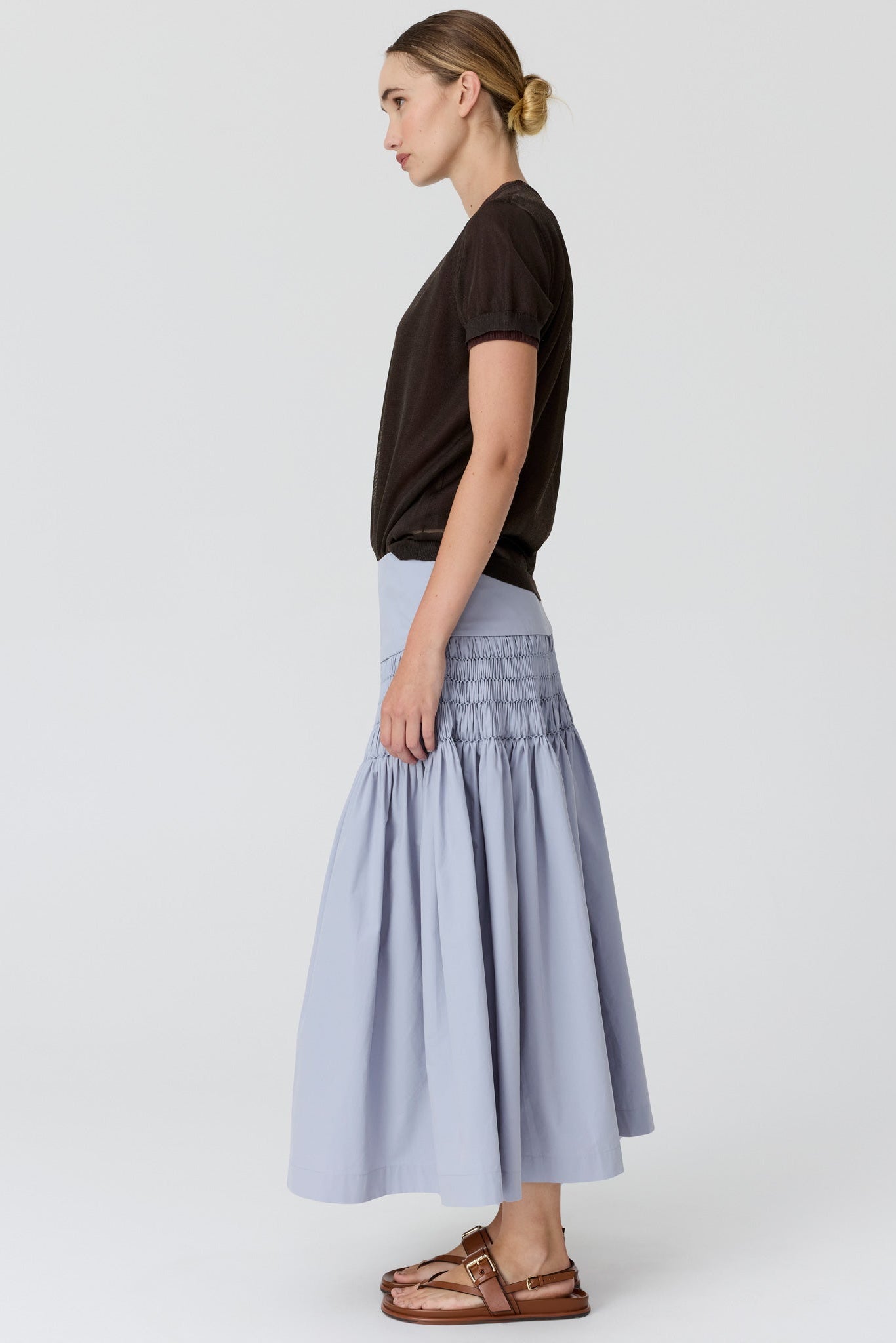 A woman shown in profile wearing a dark brown short-sleeve top and a light periwinkle blue pleated midi skirt, paired with brown leather sandals, against a plain light gray studio background.