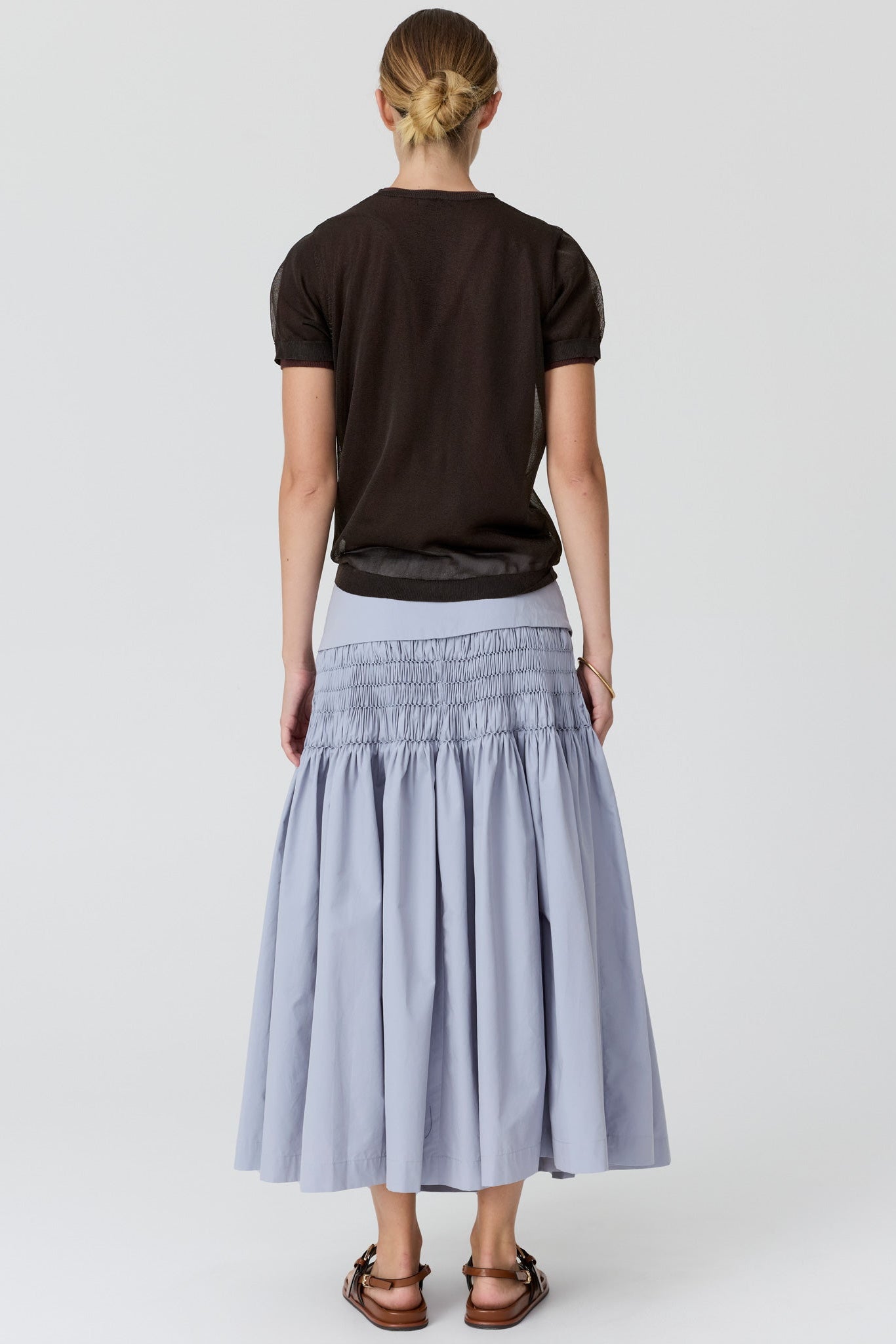 A woman viewed from the back wearing a dark brown short-sleeve t-shirt and a light blue pleated midi skirt with a smocked waistband, paired with brown sandals, against a light gray studio background.