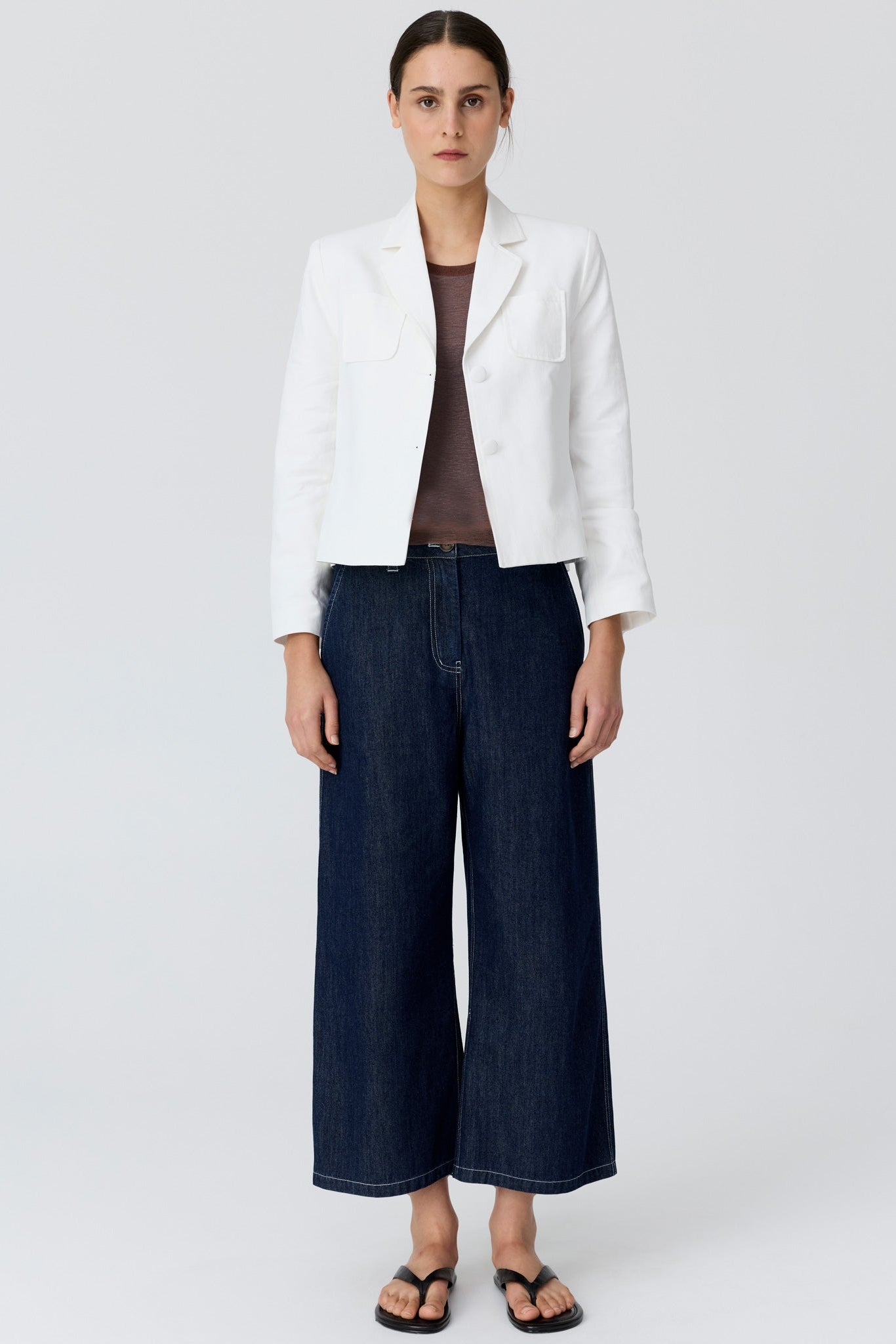 Woman shown from the back wearing a cropped white blazer and dark denim wide leg pants and sandals on a grey studio background. 