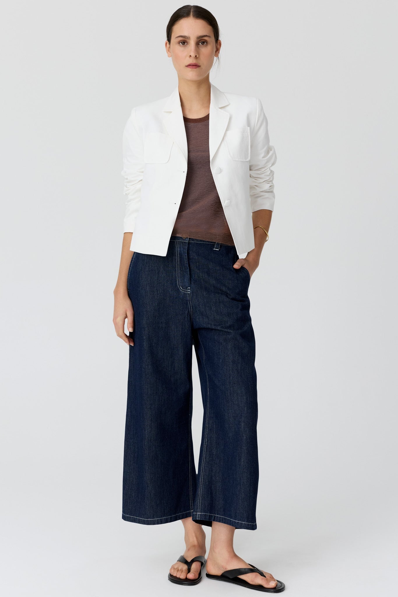 Woman shown from the back wearing a cropped white blazer and dark denim wide leg pants and sandals on a grey studio background. 