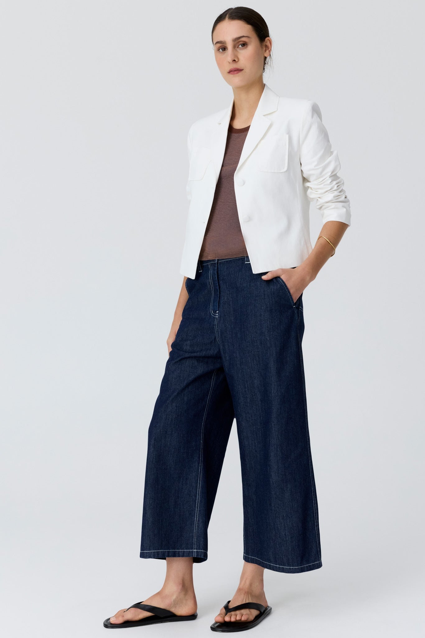 Woman shown from the side angle wearing a cropped white blazer with a tan top underneath and dark denim wide leg pants and sandals on a grey studio background. 