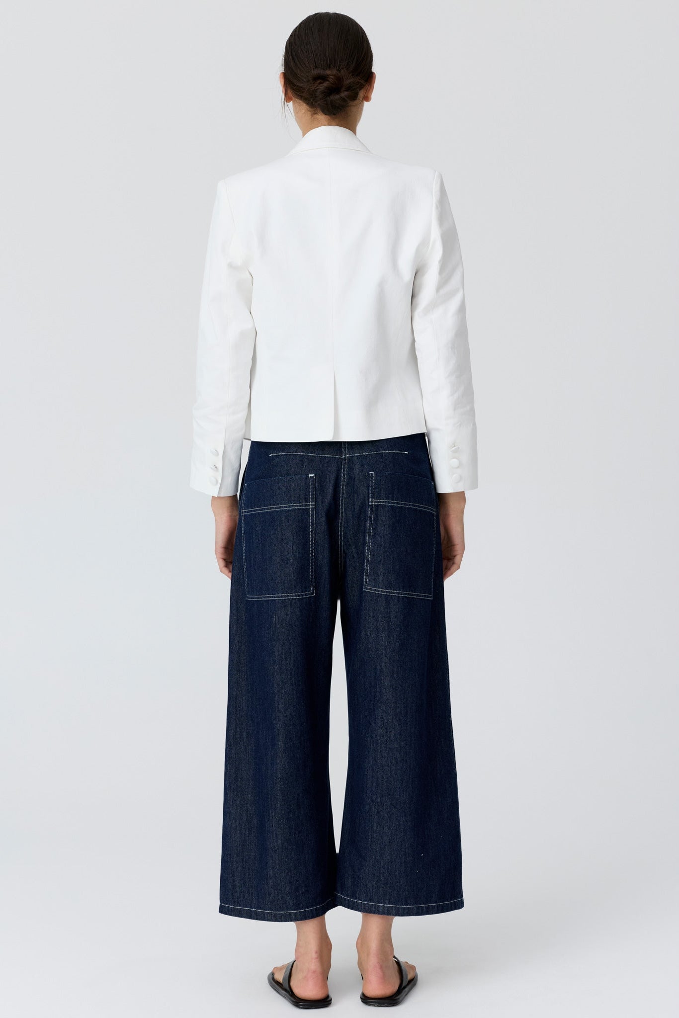 Woman shown from the back wearing a cropped white blazer and dark denim wide leg pants and sandals on a grey studio background. 