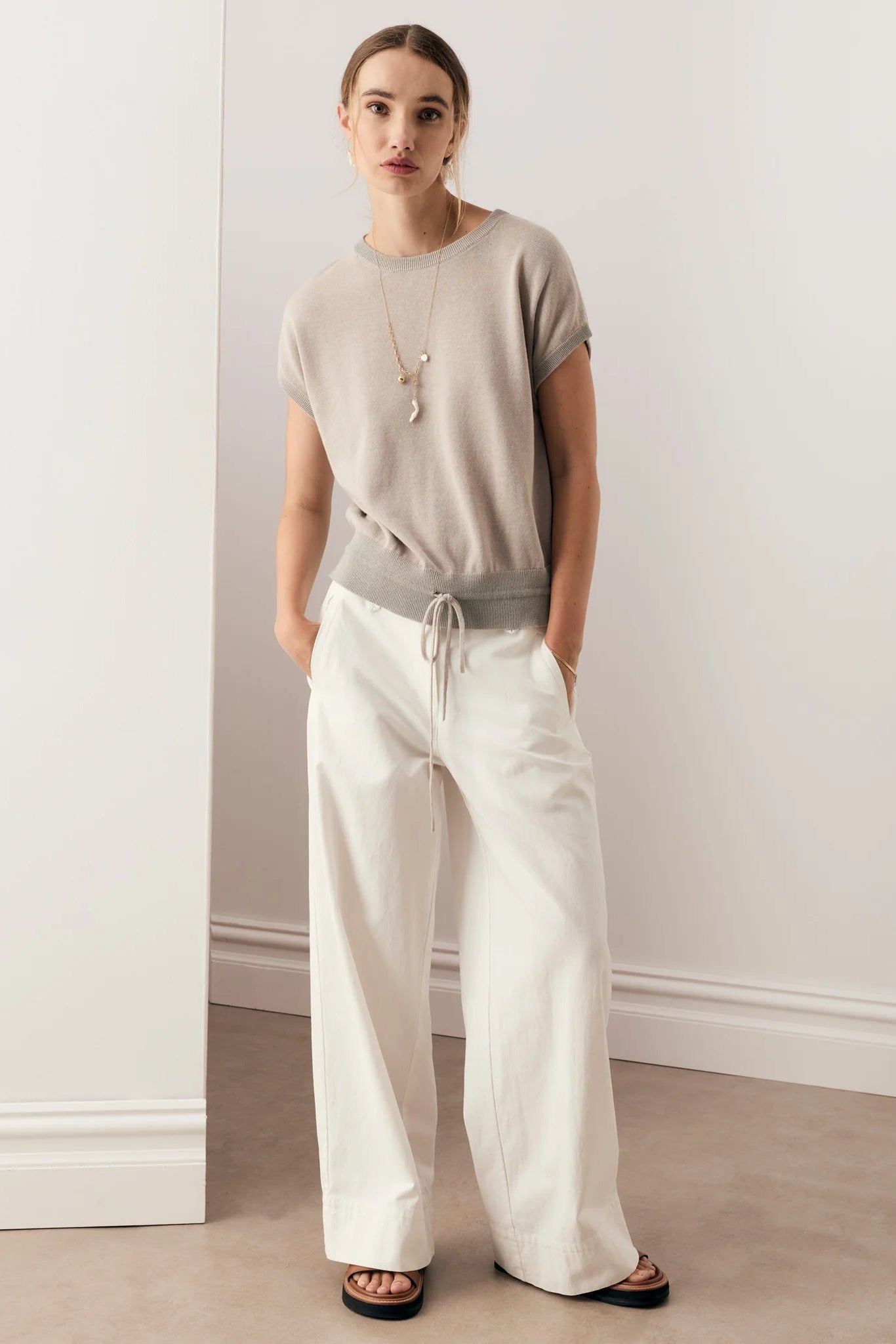 Model wearing a tan top with long necklace and white wide leg pants and sandals in a hallway setting. 