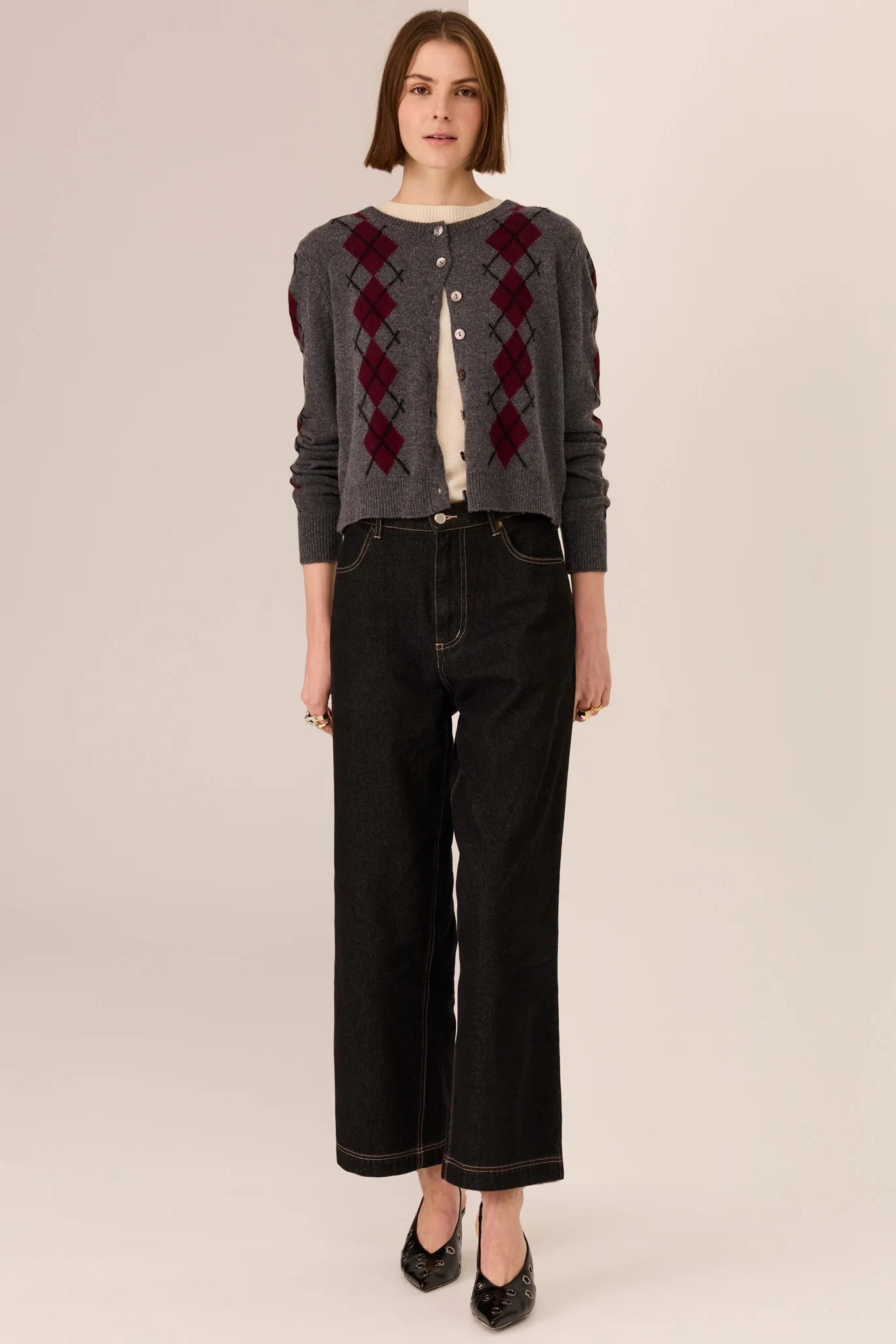 Woman wearing a gray argyle cardigan with a beige top and black pants on a plain background