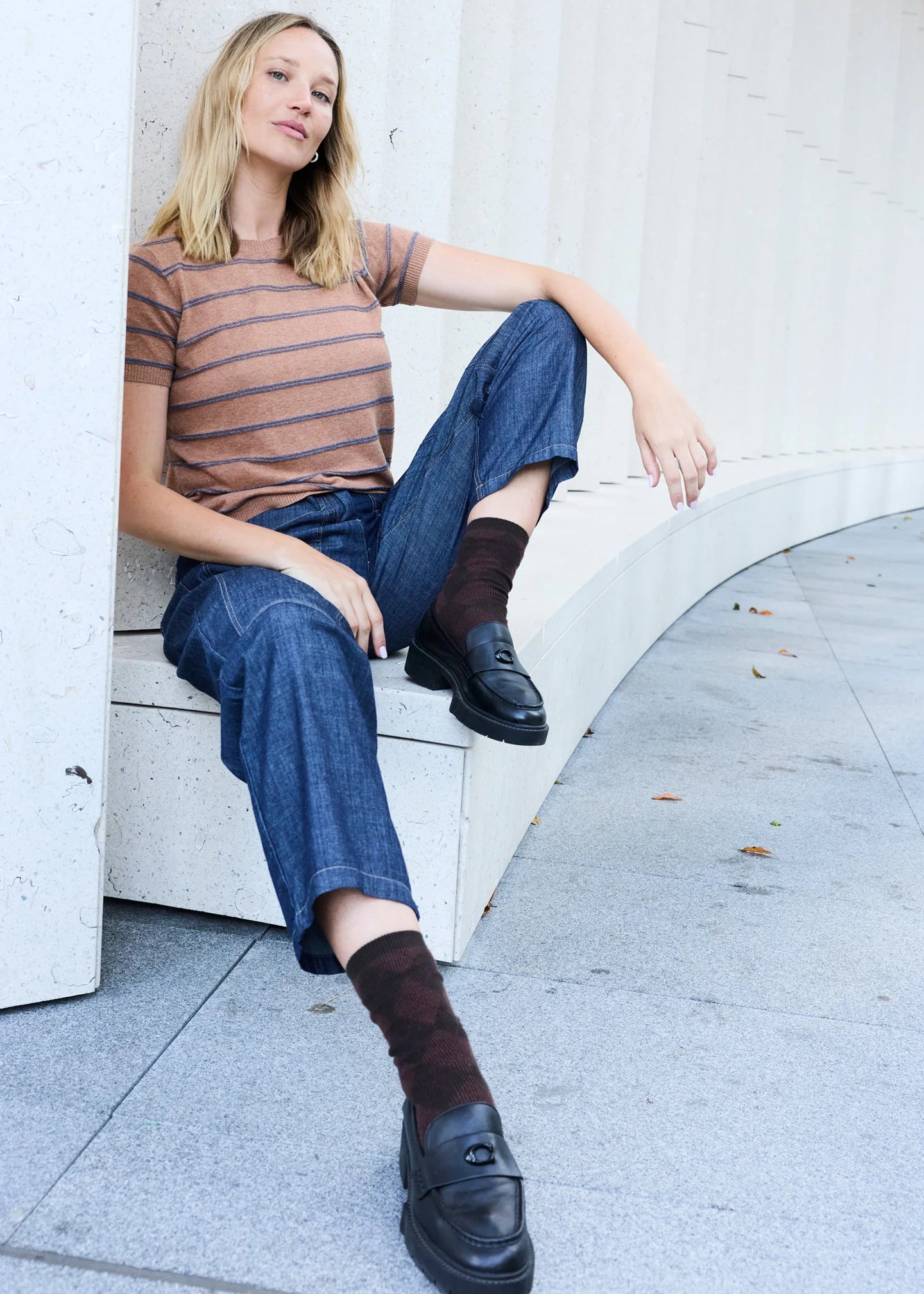 Woman sitting on a white bench wearing a striped top blue denim pants, and dark shoes. 