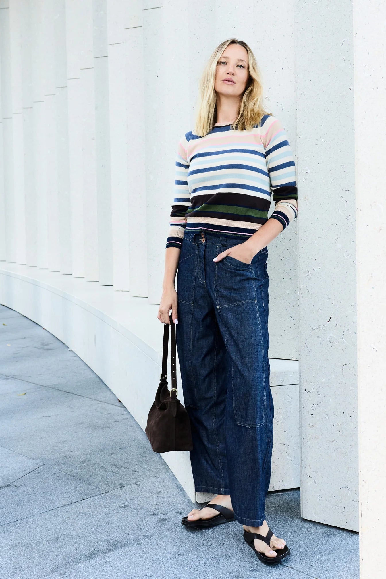 Woman wearing a striped sweater and blue jeans standing against a white wall.