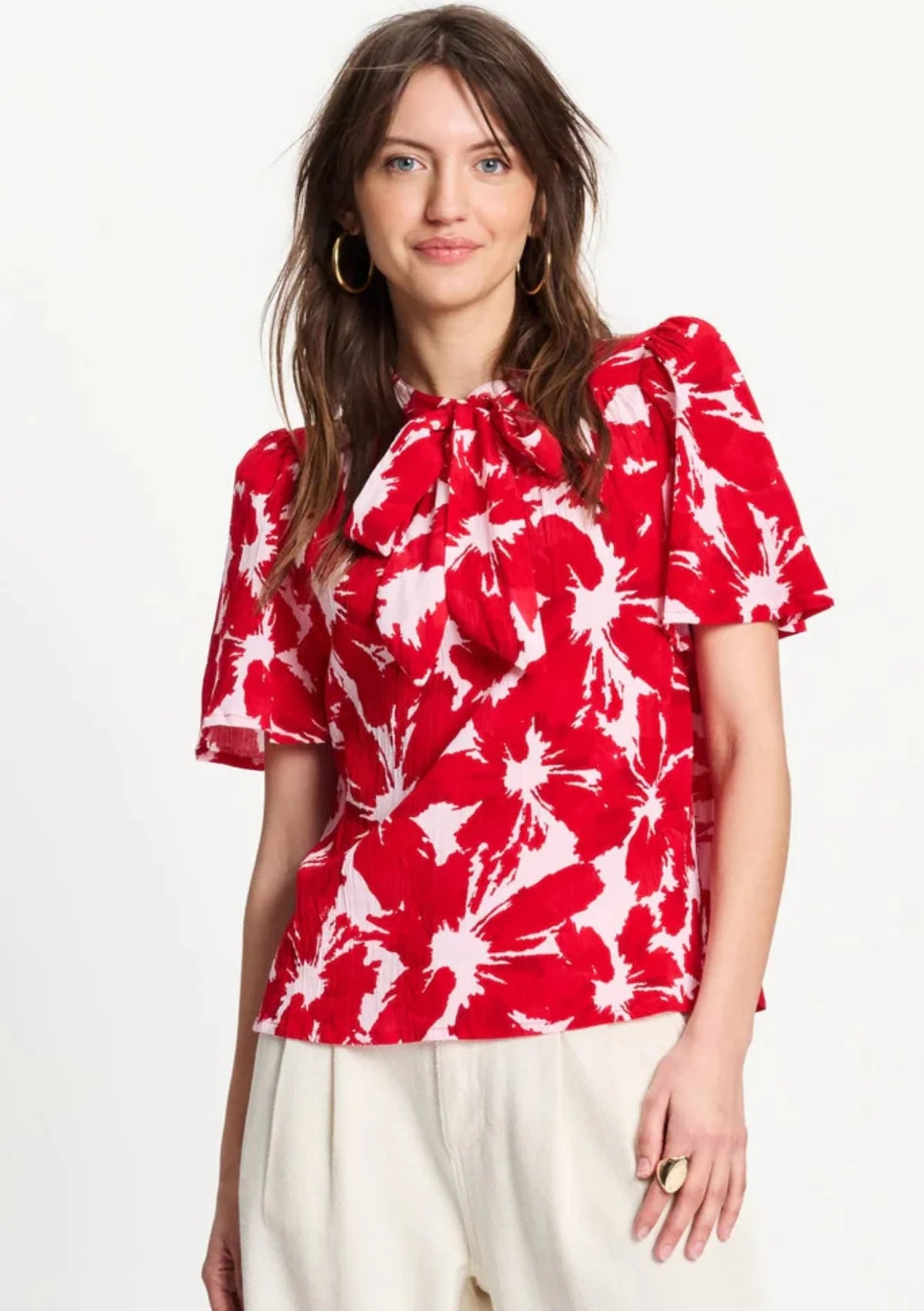 A woman wearing a red and white abstract floral print blouse with puff sleeves and a ruffled tie-neck, paired with cream trousers, against a plain white background.
