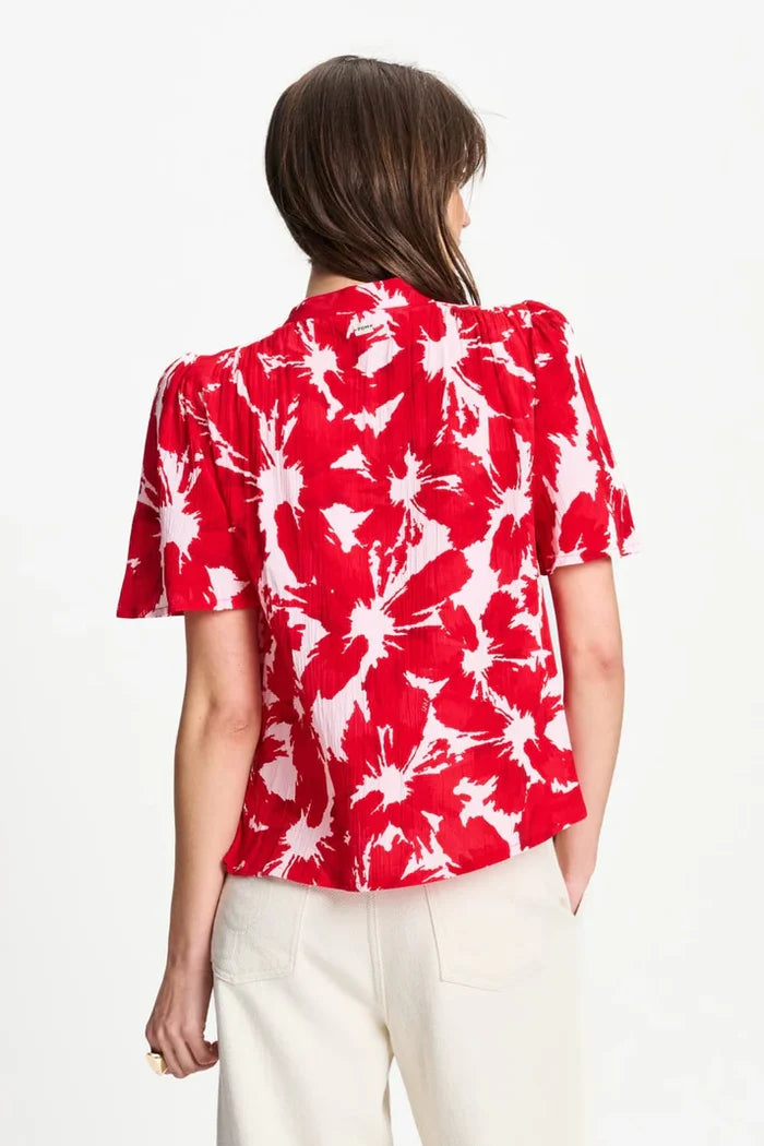 A woman shown from the back wearing a red and white abstract floral print blouse with puff sleeves and a ruffled tie-neck, paired with cream trousers, against a plain white background.