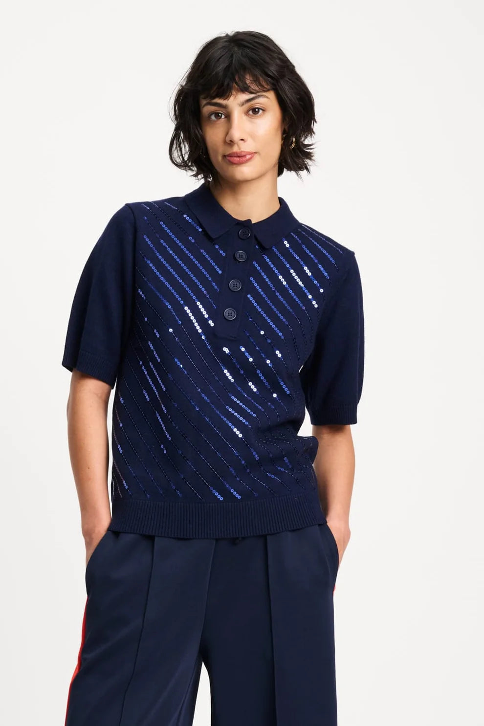 A model wearing a navy blue short-sleeve knit polo shirt with diagonal blue and silver sequin stripes across the front, paired with navy trousers, against a light gray studio background.