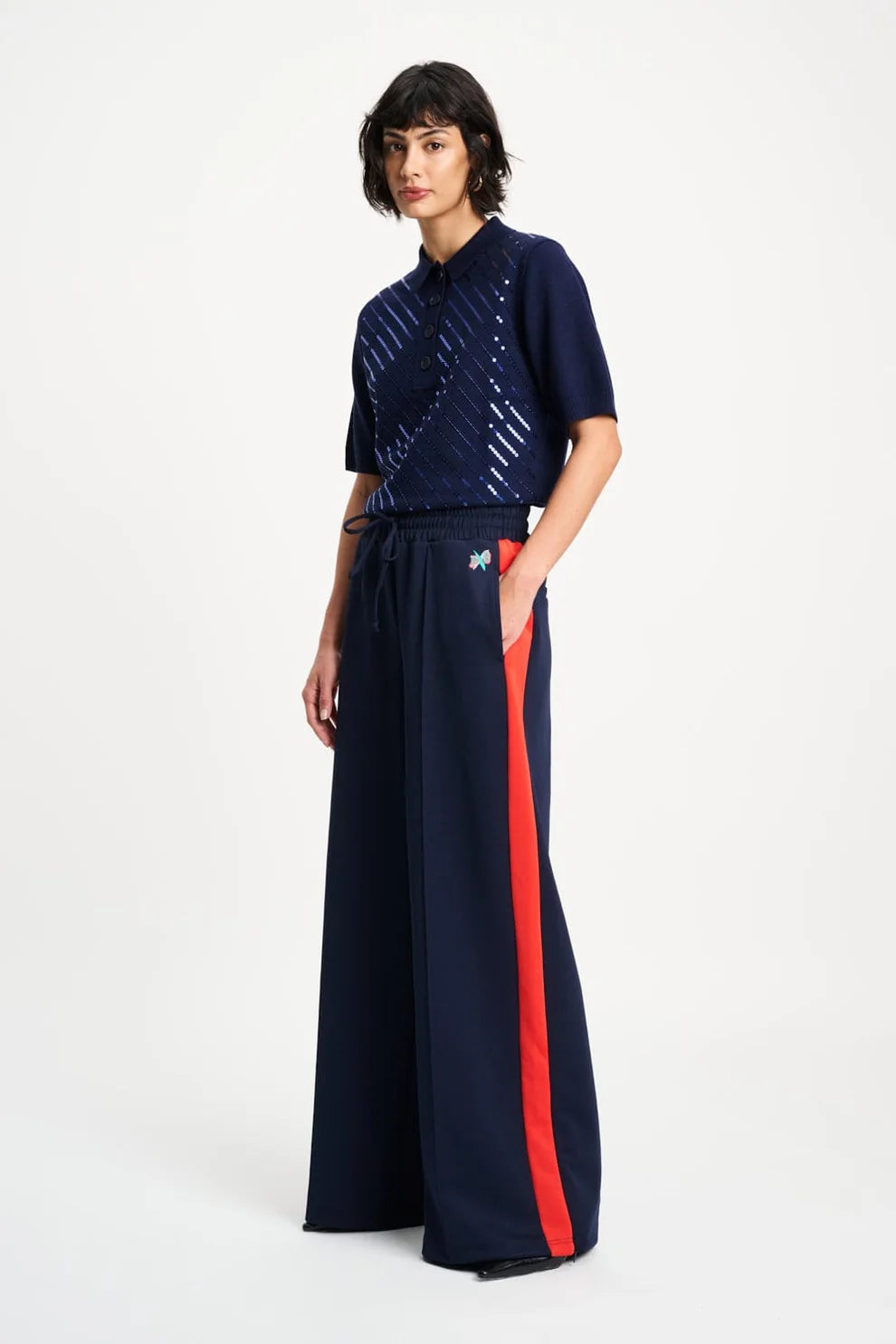 Woman wearing a navy short sleeve sequinned polo and navy pants with a red stripe on a white background