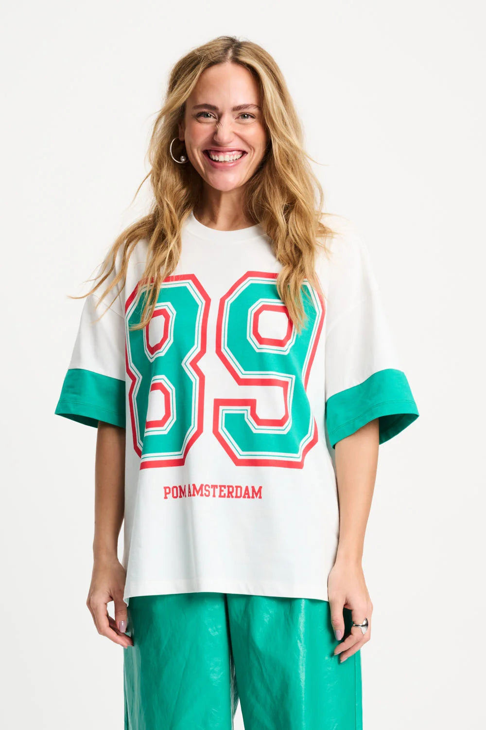 A smiling woman wearing a white oversized graphic T-shirt with large green and red outlined '89' on the front, green sleeve cuffs, and 'PON LMSTERDAM' printed below, paired with shiny green pants against a light gray studio background.