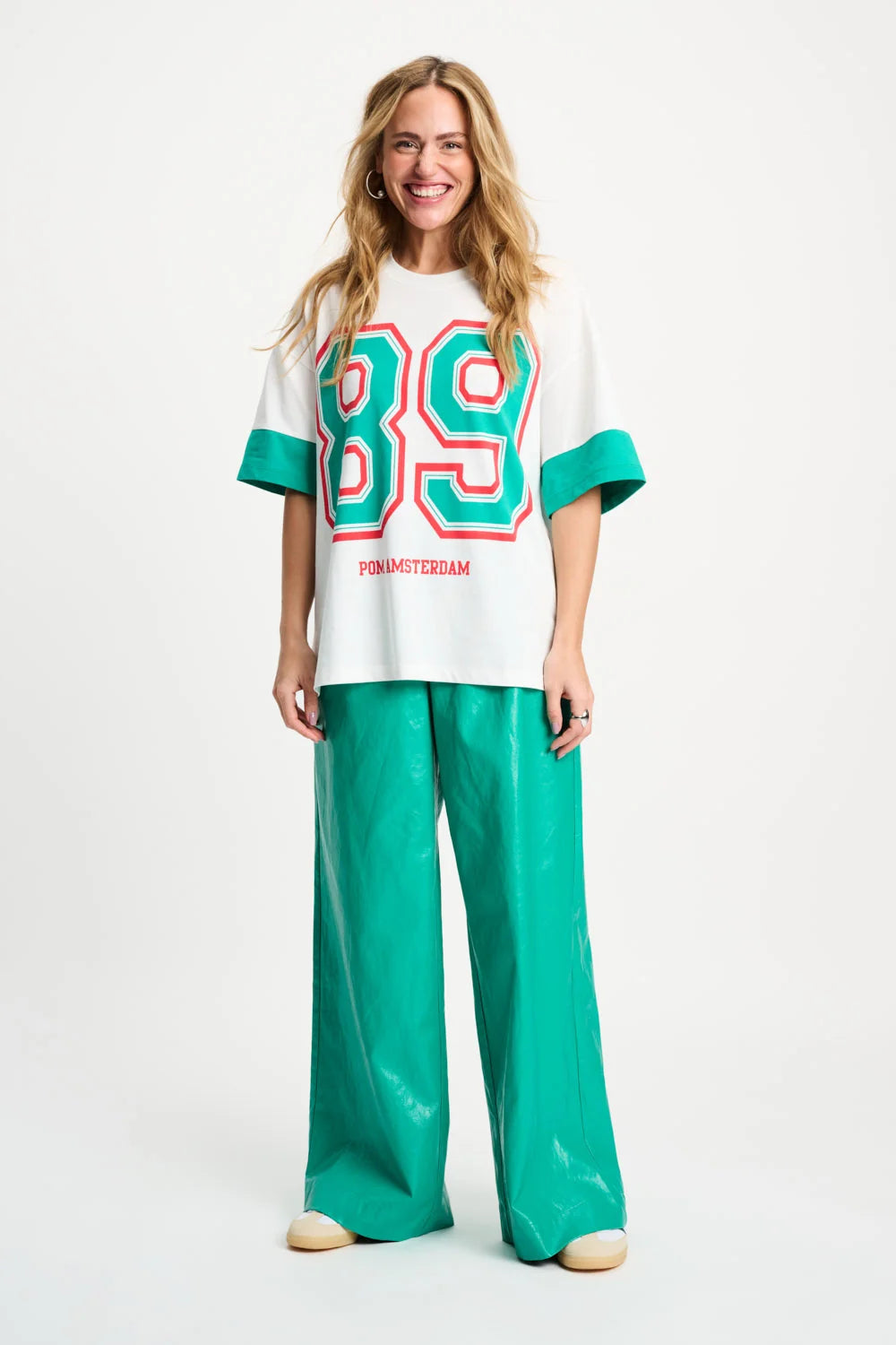 A woman modeling a white oversized t-shirt with green and red '89' graphics and 'HONOURABLE' text, paired with shiny green wide-leg pants, against a plain white studio background.