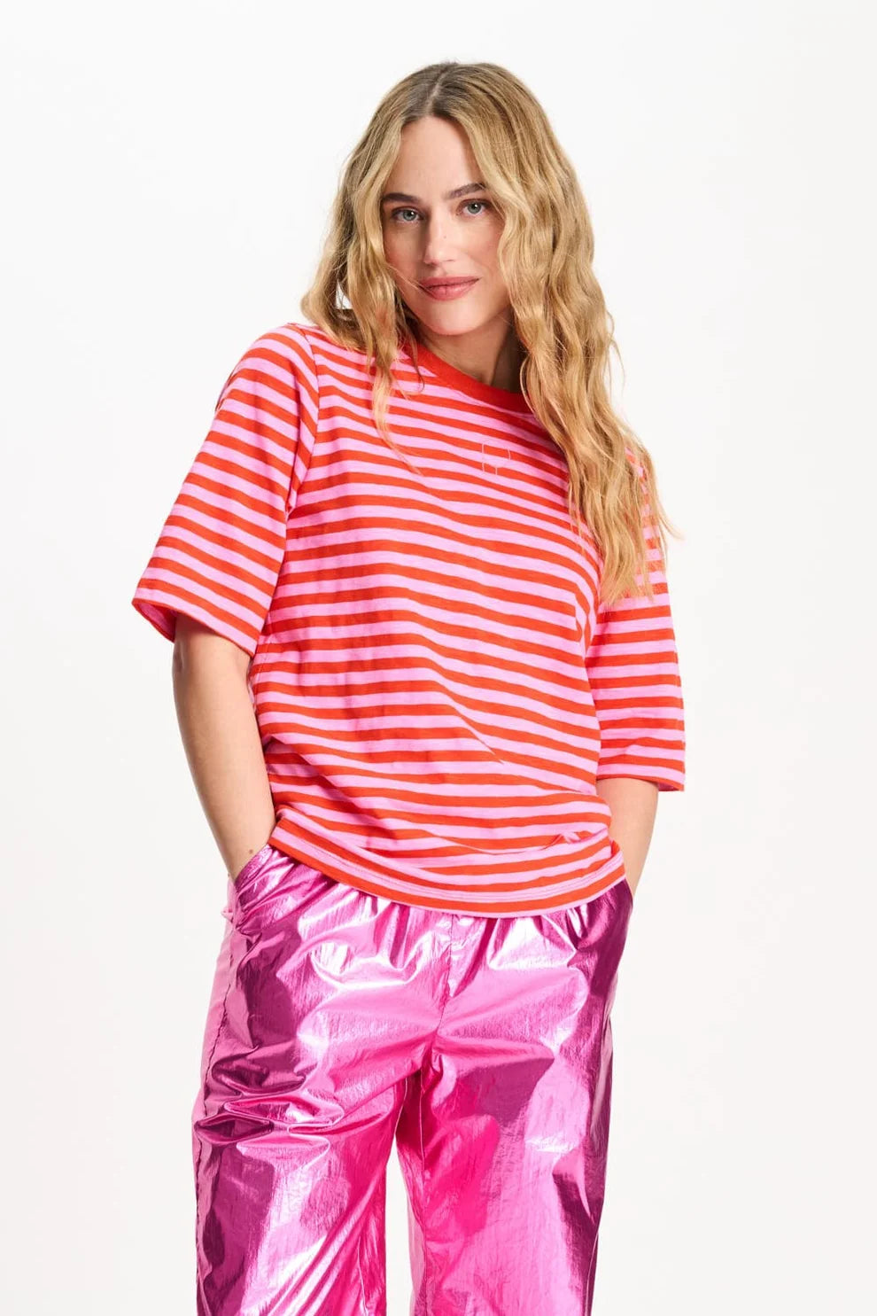 A woman modeling a orange and pink  horizontally striped short-sleeve t-shirt paired with shiny metallic pink pants, set against a plain white studio background.