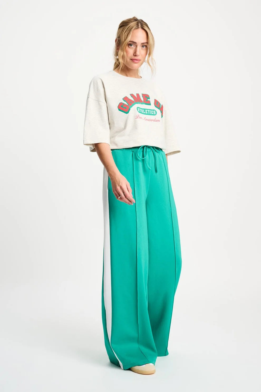 Woman wearing a white oversized shirt with text and teal pants with a white stripe on a white background