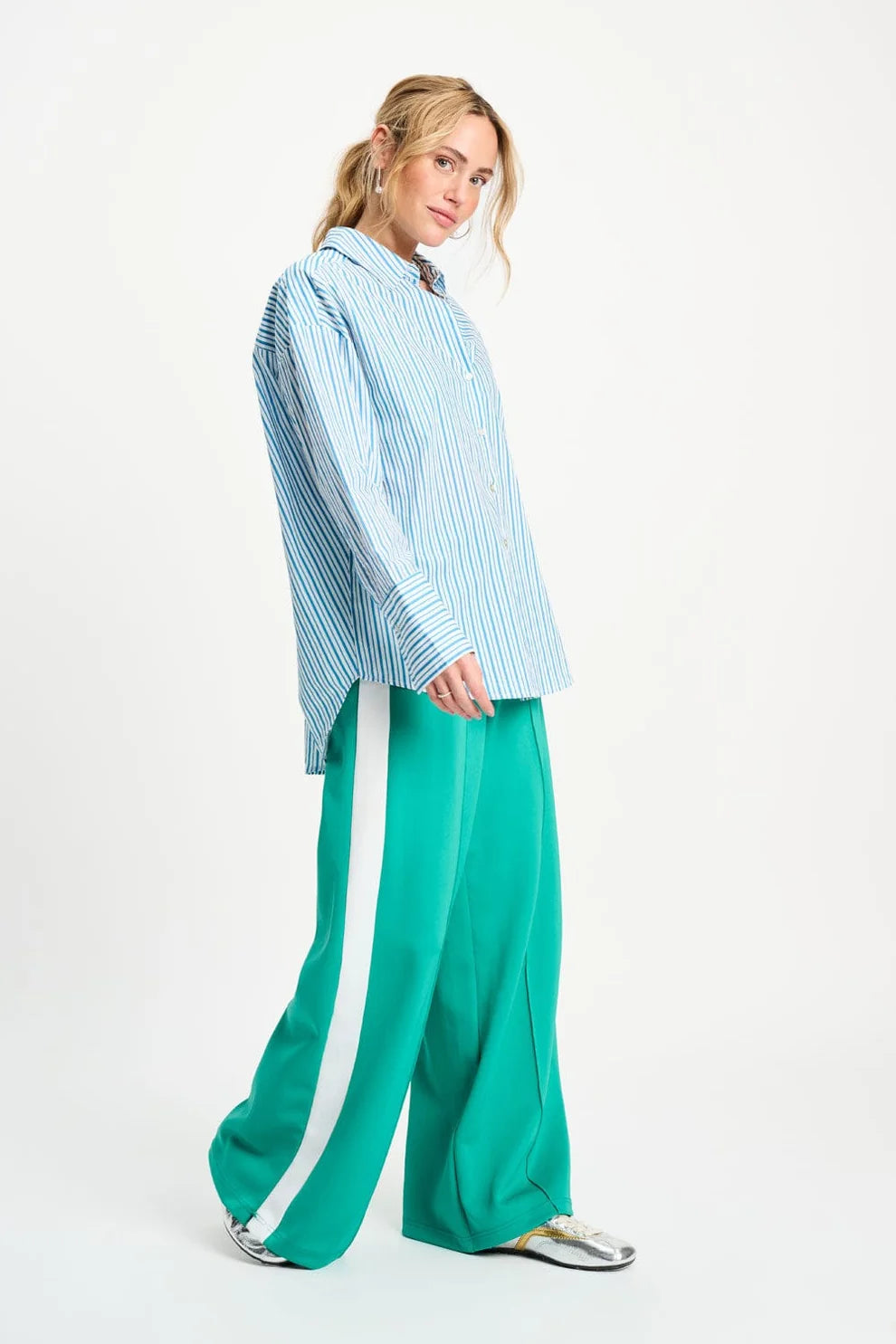 A woman modeling a light blue and white vertically striped button-up shirt paired with bright green wide-leg pants featuring a white side stripe, set against a plain white studio background.