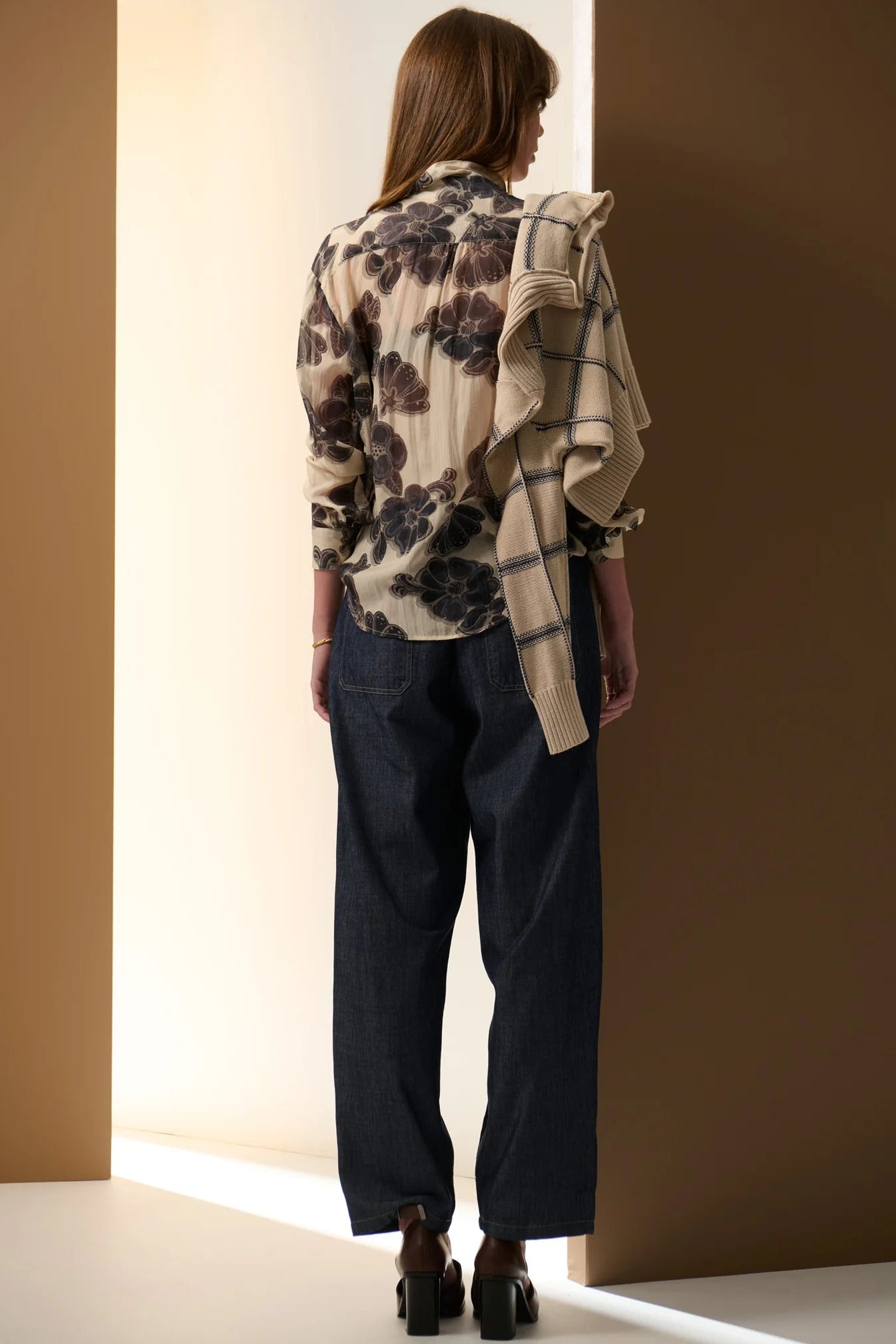 Person wearing a floral shirt and dark pants with a plaid scarf, standing against a neutral wall.