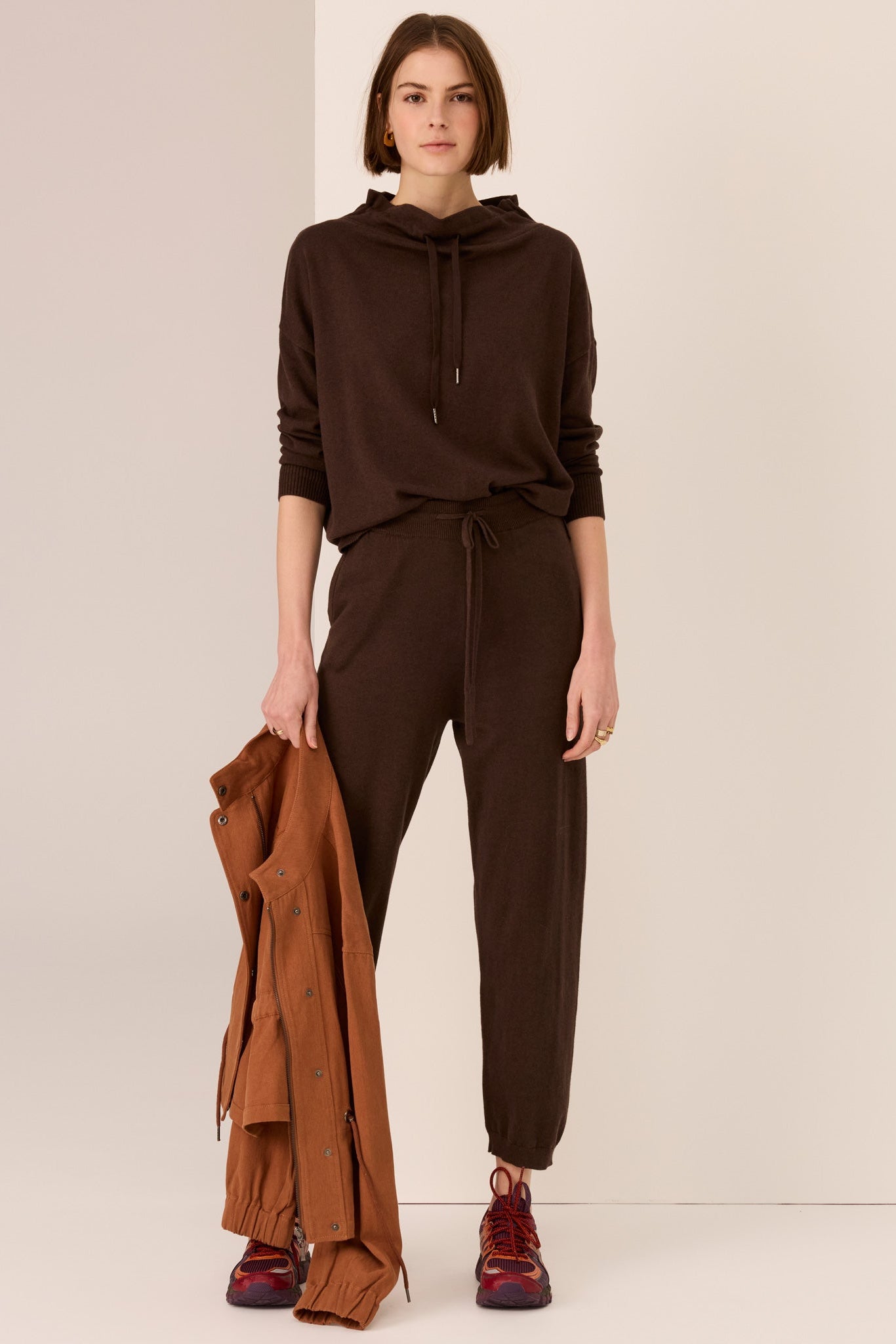 Woman wearing a brown outfit with sweater with drawstring and matching pant holdiing a tan jacket on a plain background. 
