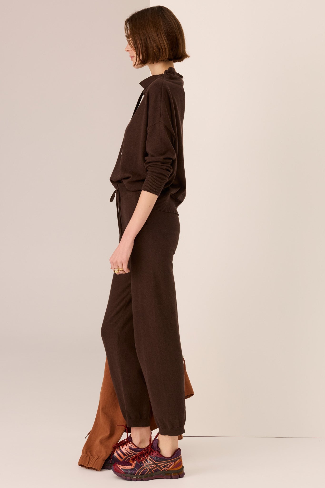 Woman wearing a brown outfit with sweater with drawstring and matching pant  on a plain background. 