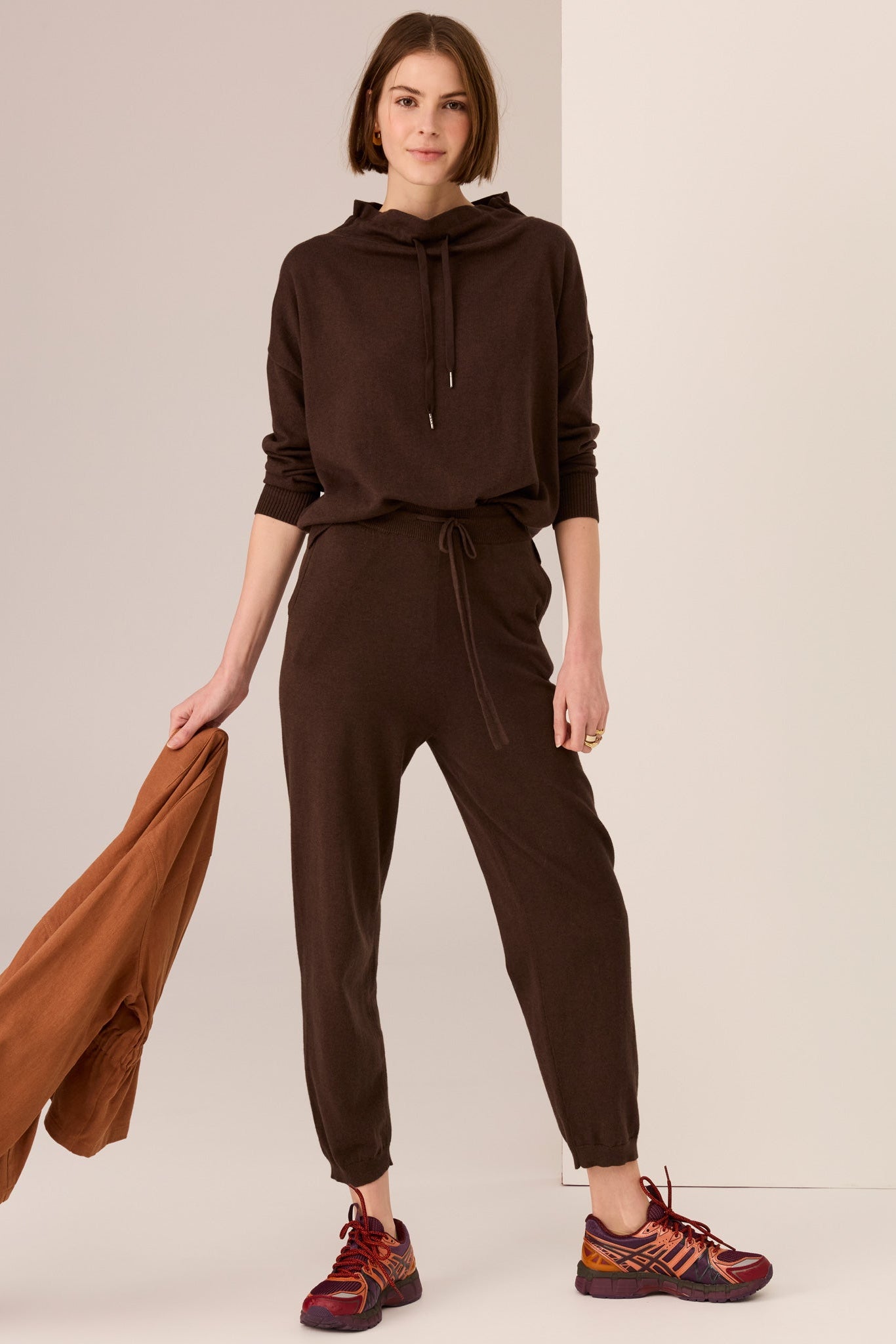Woman wearing a brown outfit with sweater with drawstring and matching pant  on a plain background. 