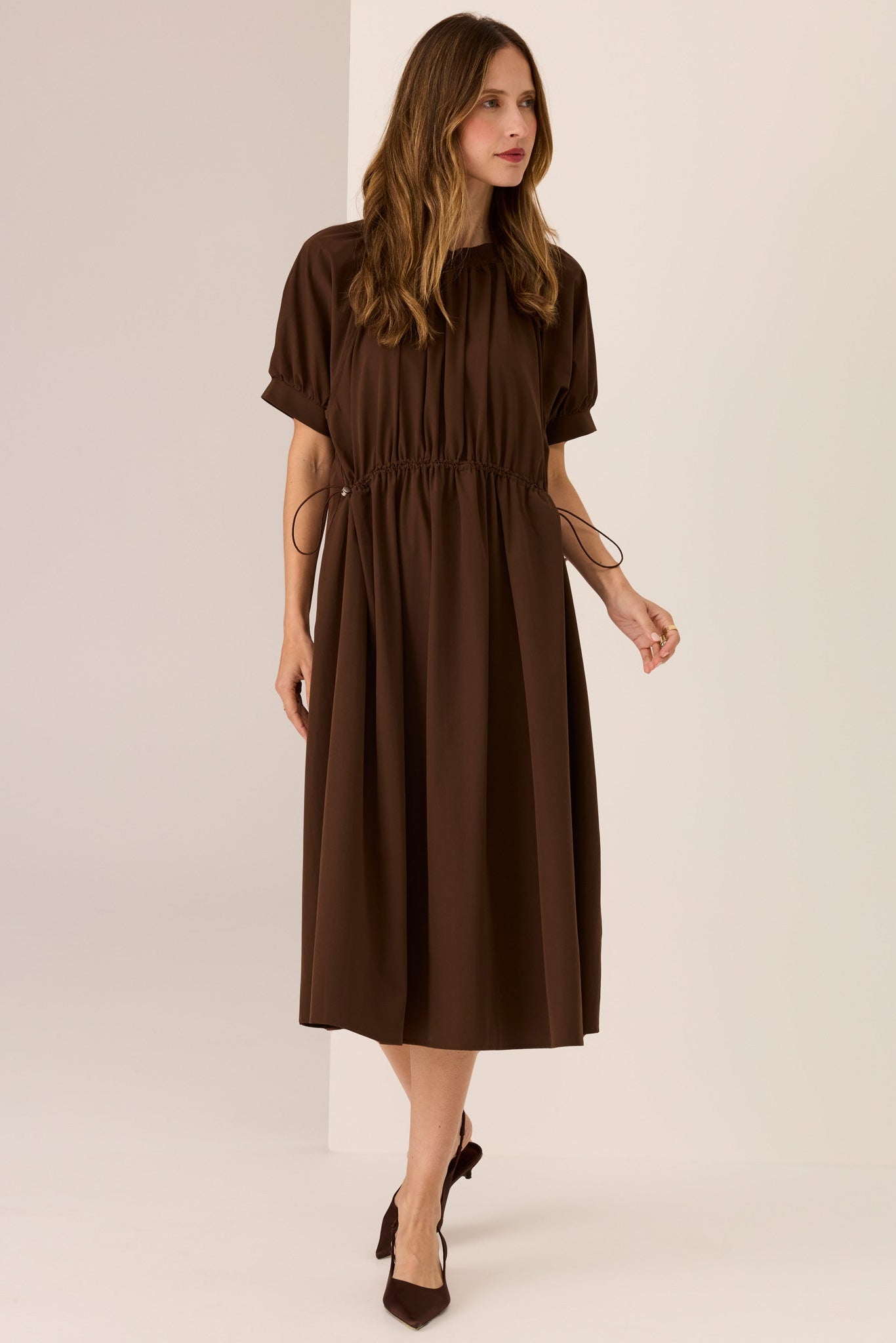 Woman wearing Chocolate brown short sleeve dress with drop waist and draw cord on a white background. 
